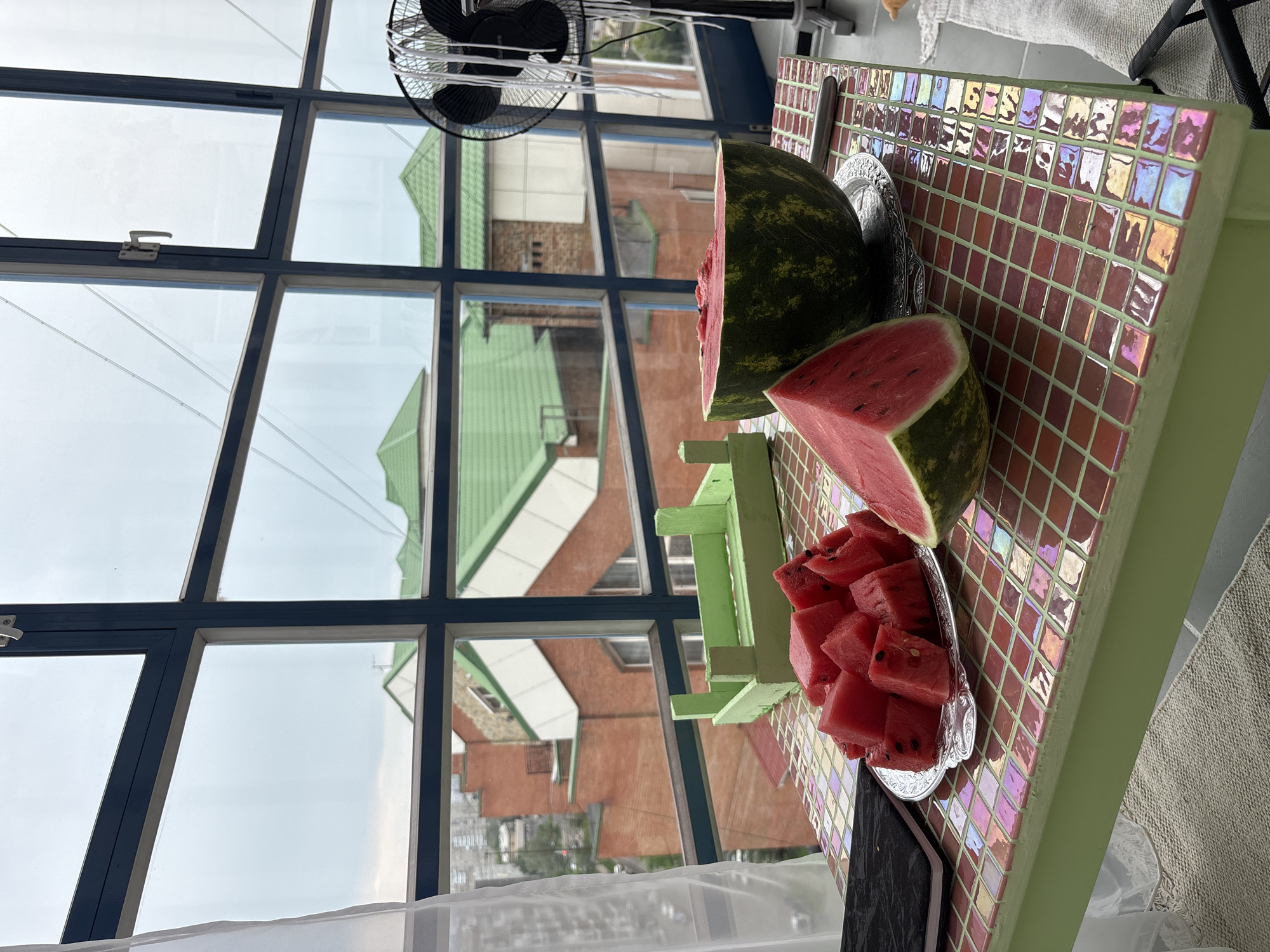 The image shows a still life of cut watermelons on a table with a tiled surface, placed in front of a large window with a view of urban buildings. The watermelons are in various states of preparation: a whole half of a watermelon is visible, along with a pile of cubed pieces on a silver platter, and a single triangular slice in the foreground. A small green wooden crate is positioned behind the cubed watermelon. The table itself is light green and appears to be covered in small, square tiles that reflect light with an iridescent quality. To the left of the table, a white sheer curtain is draped. In the background, through the window with blue frames, are brick buildings with green roofs, and what appears to be a city skyline under an overcast sky. A black oscillating fan stands to the right of the scene. The overall setting suggests an indoor space with a view overlooking a city. The location is identified as Krasnoyarsk, Russia.