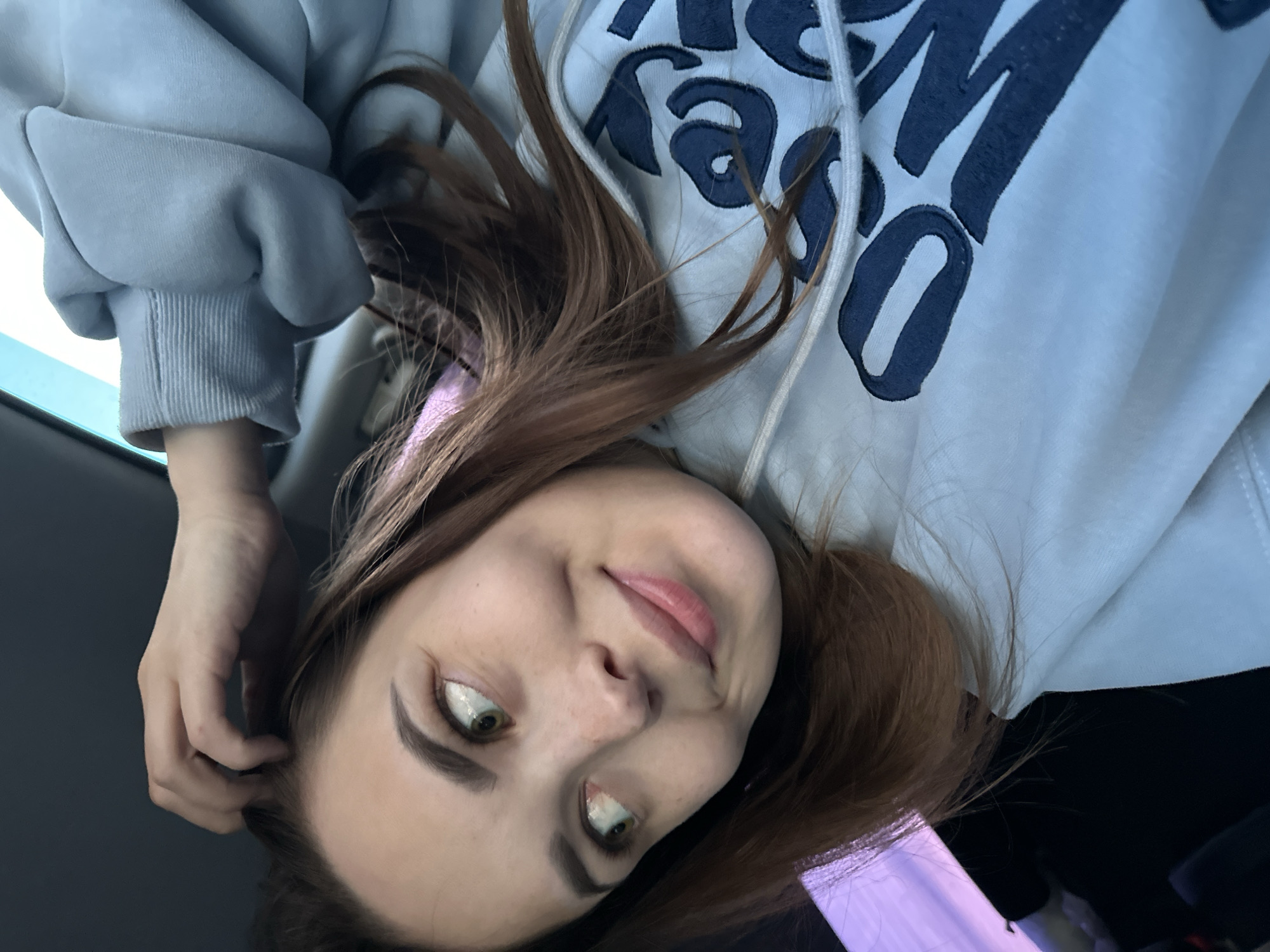 A woman is seen in a selfie, oriented upside down. She has light skin, brown hair, and green eyes. She is wearing a light-colored hoodie with dark blue lettering. Her hand is near her head, possibly touching her hair. The background is dark and indistinct, though some purple-ish light is visible. There is no explicit evidence to confirm the location.