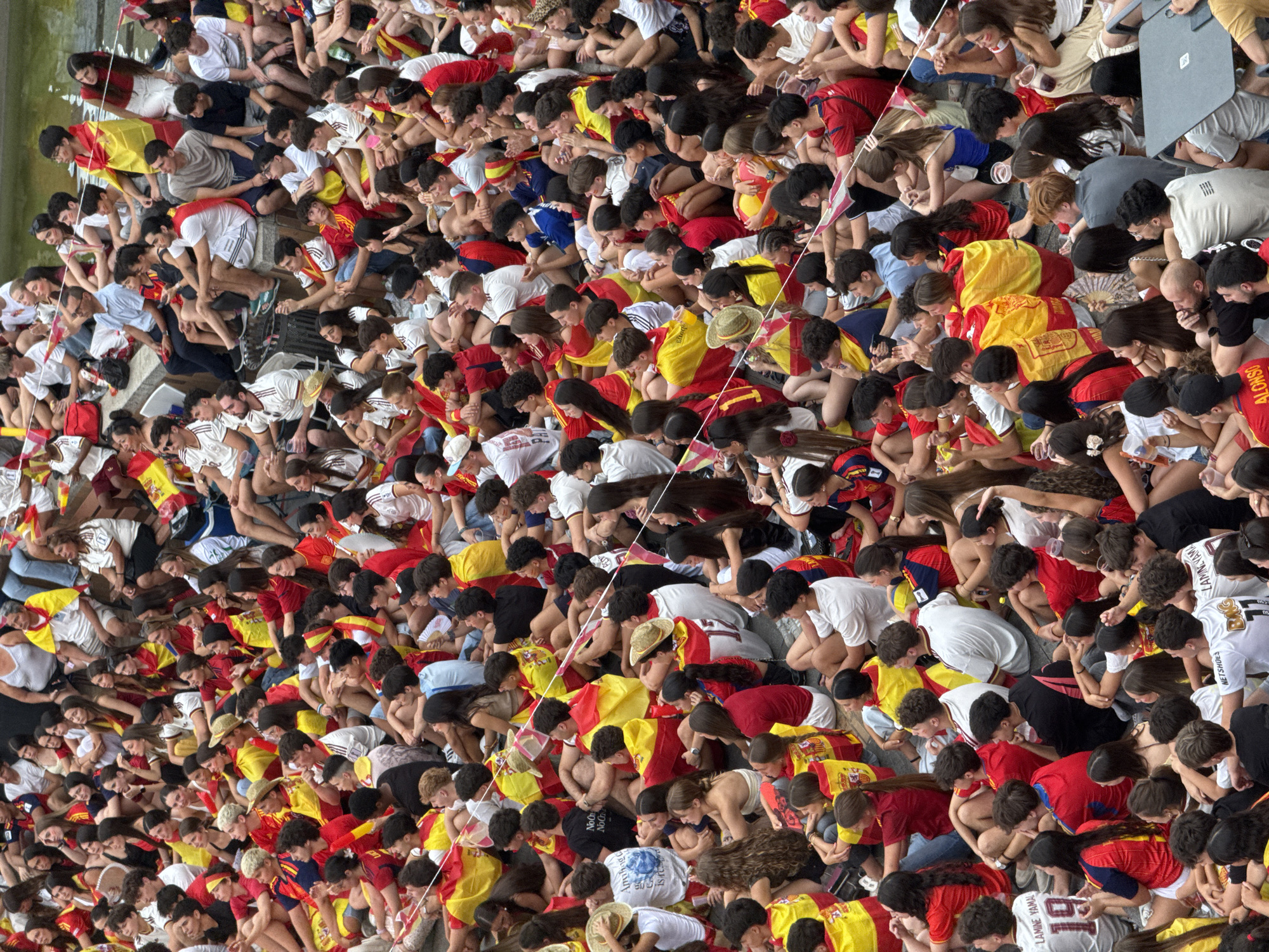 The image captures a crowd of people, predominantly young adults, gathered outdoors in a public space, likely a plaza or street, in Las Rozas de Madrid, Spain. The atmosphere appears festive and engaged, with many individuals wearing Spain's national colors of red and yellow, often in the form of flags, scarves, or t-shirts. Some people are seated on the ground, while others are standing or in smaller groups, suggesting they are watching an event or gathering together. The overall scene indicates a sense of community and shared interest, possibly related to a sporting event or a cultural celebration. The background is indistinct but suggests an urban setting, with paved ground and hints of surrounding structures or greenery. The lighting suggests it is daytime, with natural light illuminating the scene.