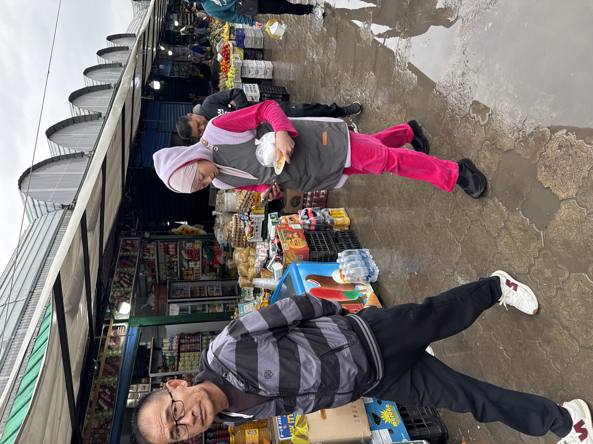 The image captures a vibrant, albeit damp, outdoor market scene in Bishkek City, Kyrgyzstan. The weather appears overcast, and recent rain is evident from the wet, puddle-strewn paved ground.
In the foreground, a man with glasses and a concerned or serious expression looks towards the viewer. He wears a grey and black horizontally striped jacket with a small "U1" logo on the chest, and dark pants. His white sneakers feature red accents and a logo resembling an inverted 'V' or 'И'. Beside him are various market goods, including bottles of oil and a carton box bearing the word "Merdon" and some Cyrillic text, possibly "Жыл-А-Тай".
In the mid-ground, a woman wears a pink outfit, a light pink hijab, and a grey apron with a small brown patch. She holds a small white plate with food wrapped in plastic and appears to be looking down at something in her hand, possibly a phone. Her black shoes are visible in a puddle. Behind her, another man in a black top is partially visible, also looking down.
The background reveals rows of market stalls extending into the distance, sheltered by a distinctive roof structure composed of multiple arching, corrugated metal sections. The stalls are laden with an assortment of products, including fresh fruits (apples, bananas, oranges), vegetables, bottled water, packaged goods, and beverages like Coca-Cola. A blue cooler with images of ice cream is also present. Text on some signs within the stalls is indistinct but appears to be product labels.
The scene depicts everyday commerce in a bustling market environment, with shoppers and vendors navigating the damp conditions.