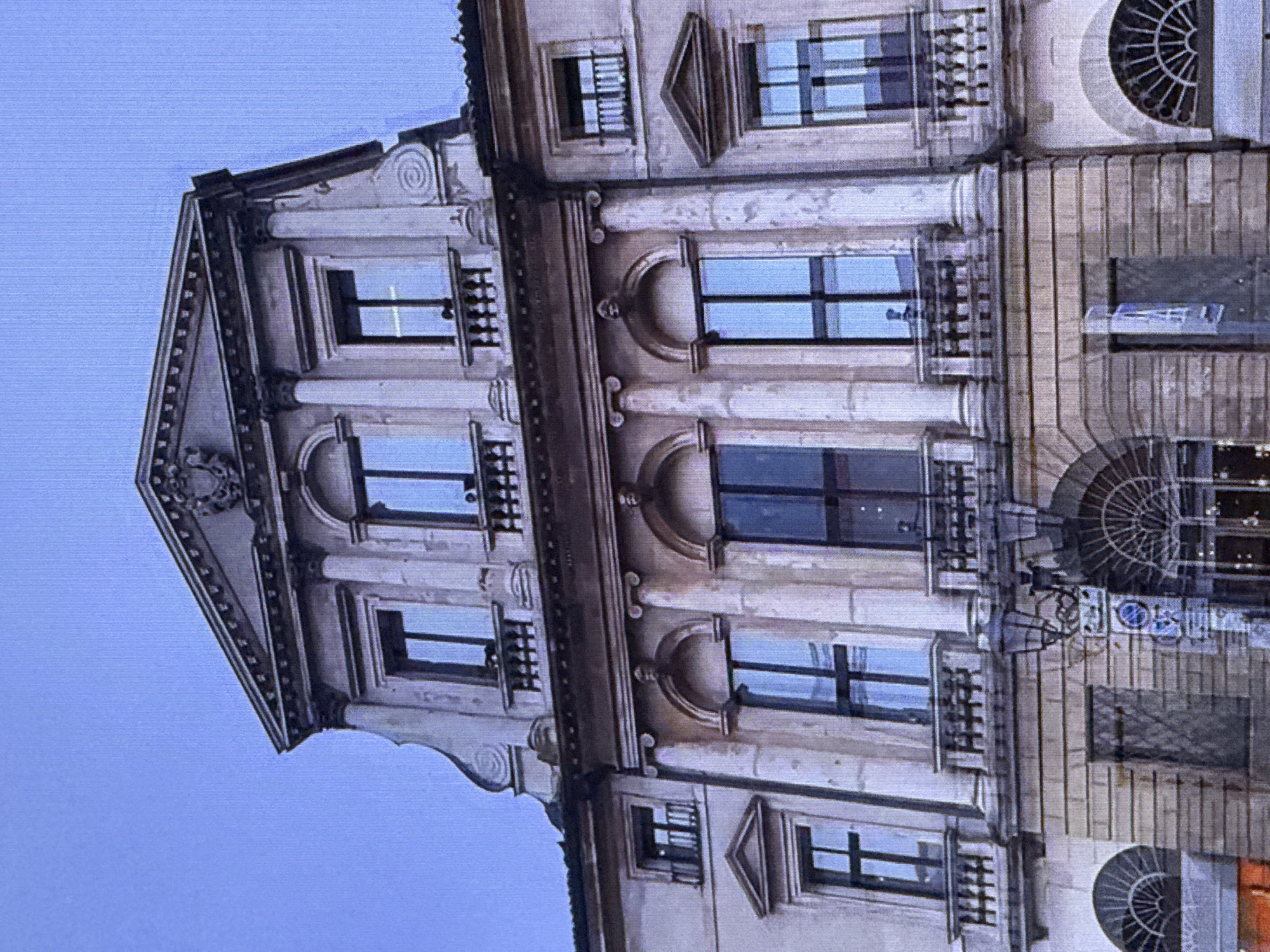 The image displays a grand, classical-style building with multiple stories and ornate architectural features. The building is constructed of stone, with a light-colored facade that shows signs of age and weathering. It features numerous windows, many of which are tall and arched, set within recessed frames and adorned with decorative moldings. Balconies with wrought-iron railings are visible on several levels. The roofline is elaborate, with a pediment at the top, decorated with a crest or coat of arms. The sky above is a clear, pale blue, suggesting daytime, though possibly early morning or late afternoon given the soft lighting.
This scene is not located in Dubai, United Arab Emirates. The architectural style is characteristic of European cities, not the modern and Islamic architectural influences typically found in Dubai.