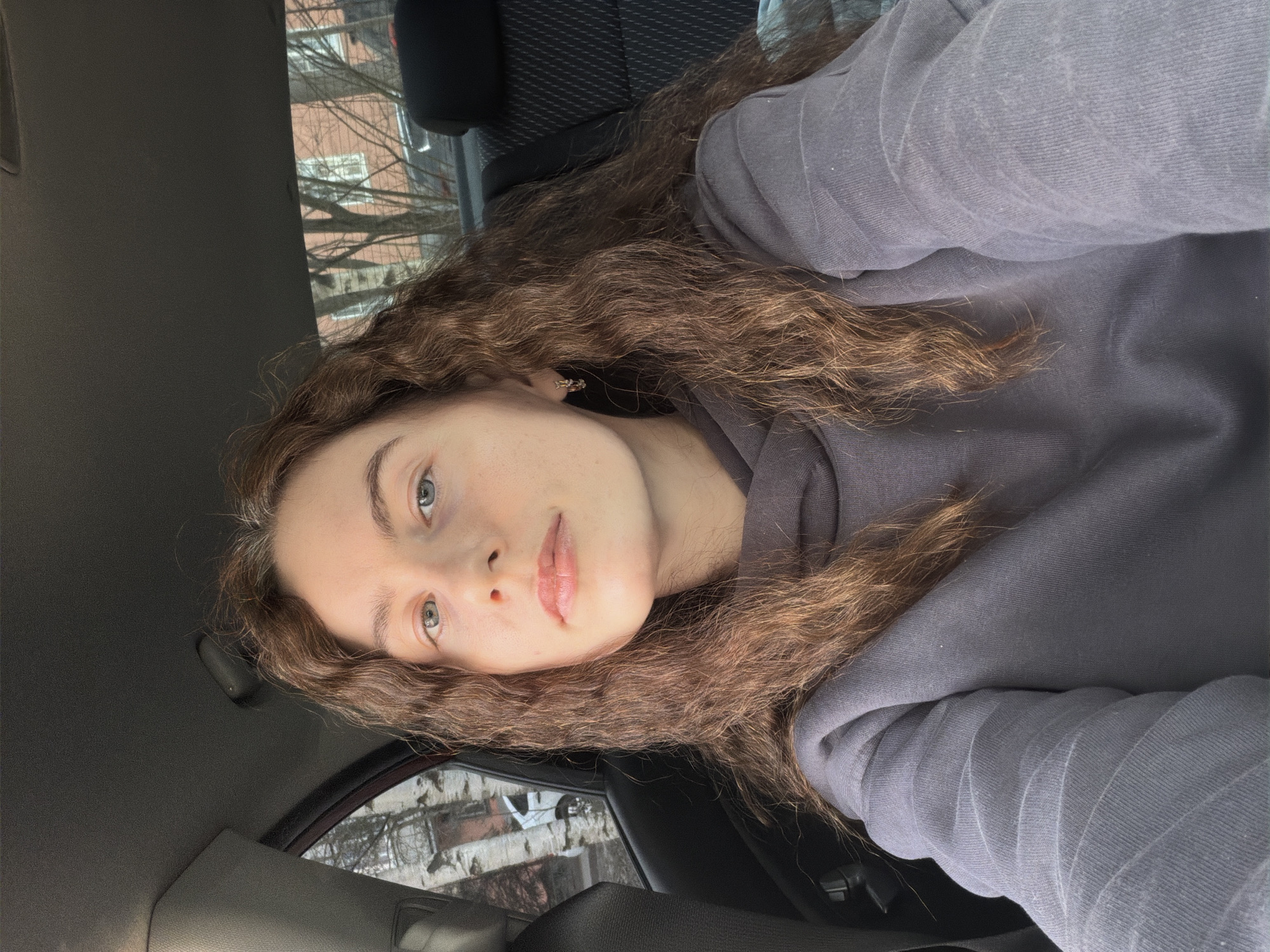 A selfie of a woman with long, wavy brown hair inside a car. She is wearing a gray hooded sweatshirt. Through the car windows, buildings and bare trees suggest a residential area. Based on the context, the location is Vsevolozhsk, Russia.