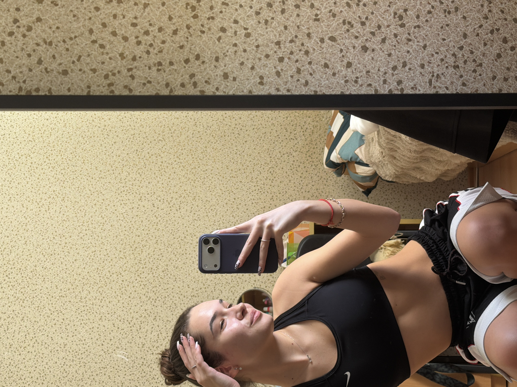 A young woman is taking a selfie in a mirror. She is wearing a black sports bra and black athletic shorts with white trim. She is holding a dark-colored cell phone up at an angle. She has long nails and a bracelet. The background features speckled wallpaper and part of a pillow with geometric patterns. The location is not discernible as Sofia, Bulgaria.