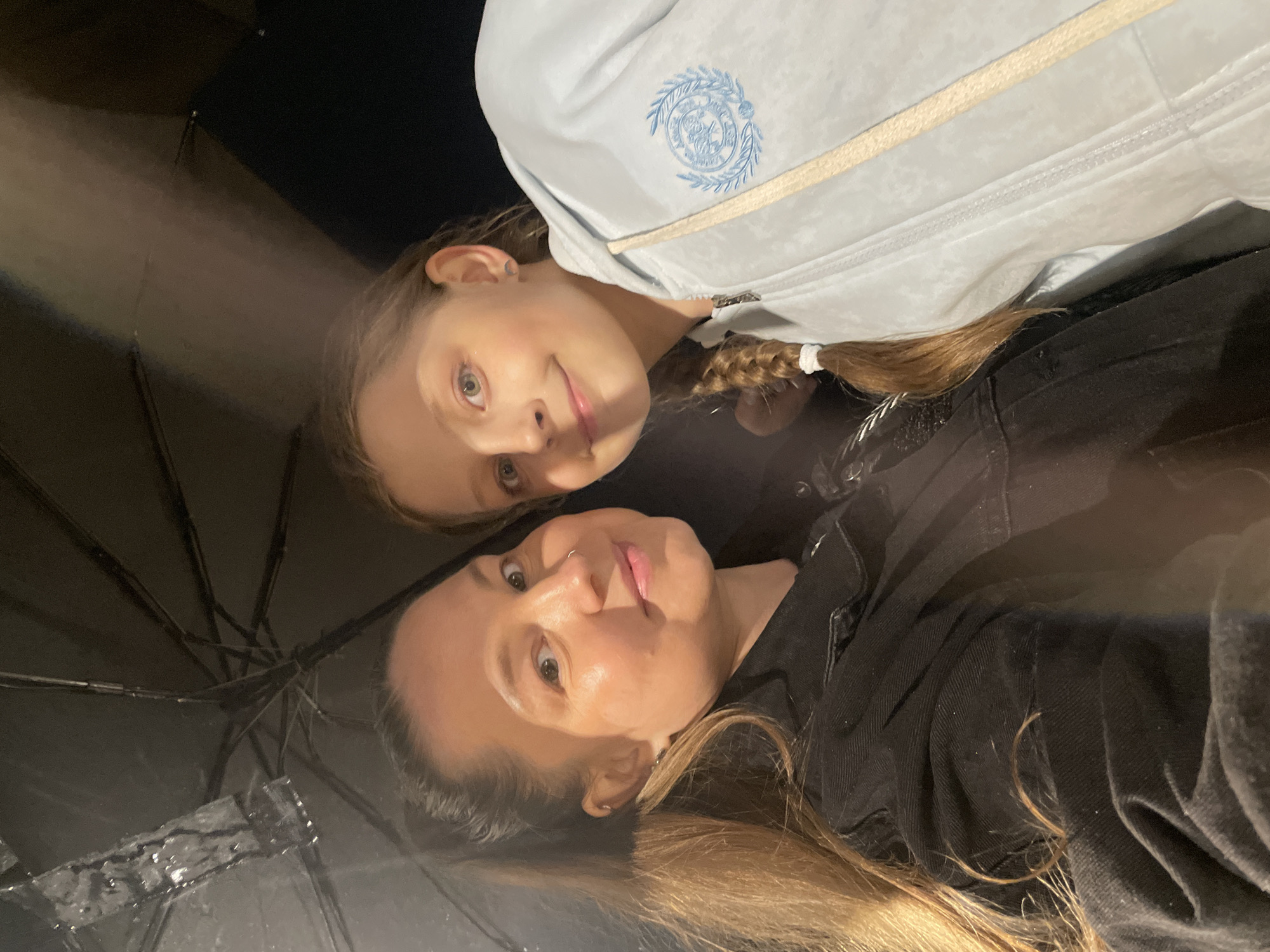 A photograph depicts two individuals, one adult and one child, positioned closely together under an open umbrella in what appears to be outdoor conditions. The image is oriented upside down. The child, positioned at the top of the frame, is fair-skinned with light hair braided and tied with a white band. The child is wearing a light blue zippered jacket with an embroidered emblem on the chest. The adult, fair-skinned with blonde hair, is wearing a black jacket or shirt. Part of the black umbrella is visible with water droplets present. The location is not identifiable as Izhevsk, Russia based on the visual information provided.