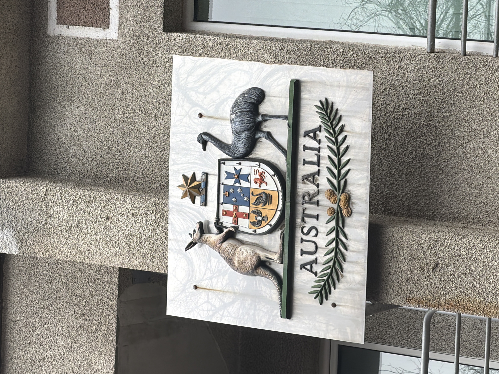 The image depicts the Australian coat of arms displayed on a building. The coat of arms features a shield divided into six sections, each representing a state of Australia, flanked by a kangaroo and an emu, and surmounted by the Commonwealth Star. The word "AUSTRALIA" is prominently displayed beside the shield. The sign is attached to a rough, grey exterior wall. The building is located in Sofia, Bulgaria, as evidenced by previous context. The image appears to be taken outdoors in daylight. A window with metal bars is visible in the background.