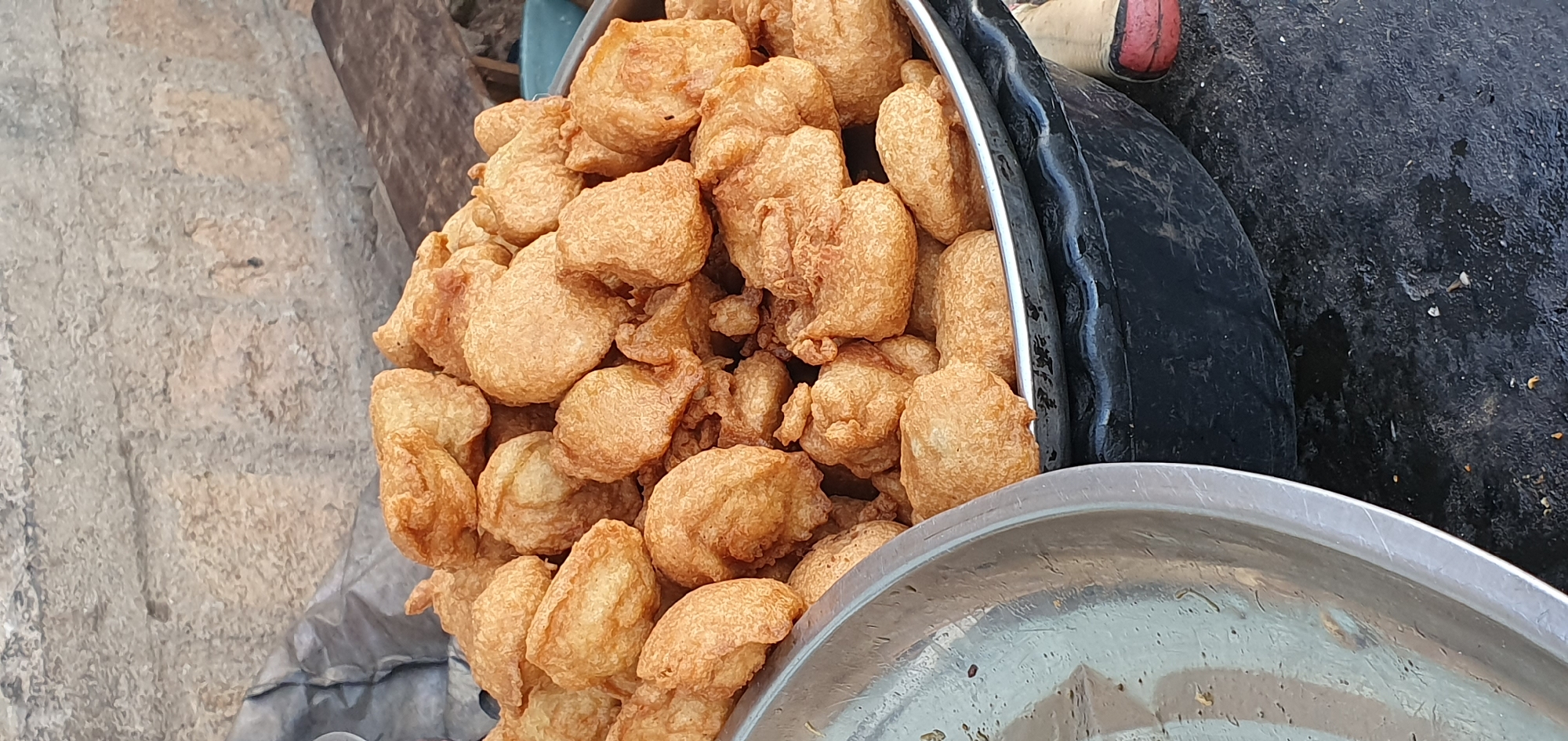 A close-up, eye-level shot shows a large pile of golden-brown, deep-fried food items, possibly fritters or dumplings, filling a dark, textured bowl. The food is unevenly shaped and appears crispy, with some pieces overlapping. To the left of the bowl, a portion of a shiny, silver bowl or dish is visible, with a textured surface and some scattered dark specks. The background is a rough, grey, stone-like surface, with a darker, crumpled material or plastic sheeting behind the food. A dark, textured rim of what appears to be a larger container or the base of the bowl holding the food is visible at the bottom of the image. The scene is outdoors, possibly a street market in Bukuru, Nigeria, given the general aesthetic of the food presentation and the textured outdoor environment.