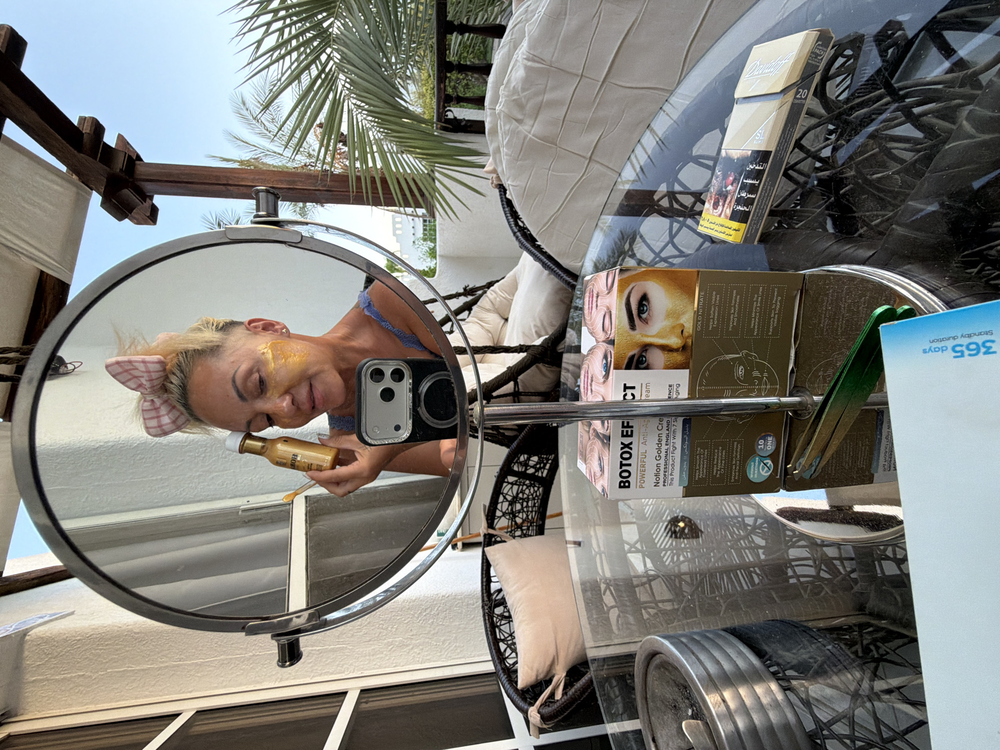 A blonde woman with a pink bow in her hair applies a golden face mask, reflected in a circular mirror. She is holding a small bottle of serum and a spatula. The mirror is placed on a table outdoors, with a view of palm trees and a clear blue sky. In the foreground, on the table, are several items: a box of "Botox Effect" cosmetic products, a pack of cigarettes, and a pair of tweezers. The woman is wearing a blue top. The setting appears to be a balcony or patio in Sharm El Sheikh, Egypt, suggested by the presence of palm trees and the architectural style. The scene captures a moment of personal grooming outdoors.