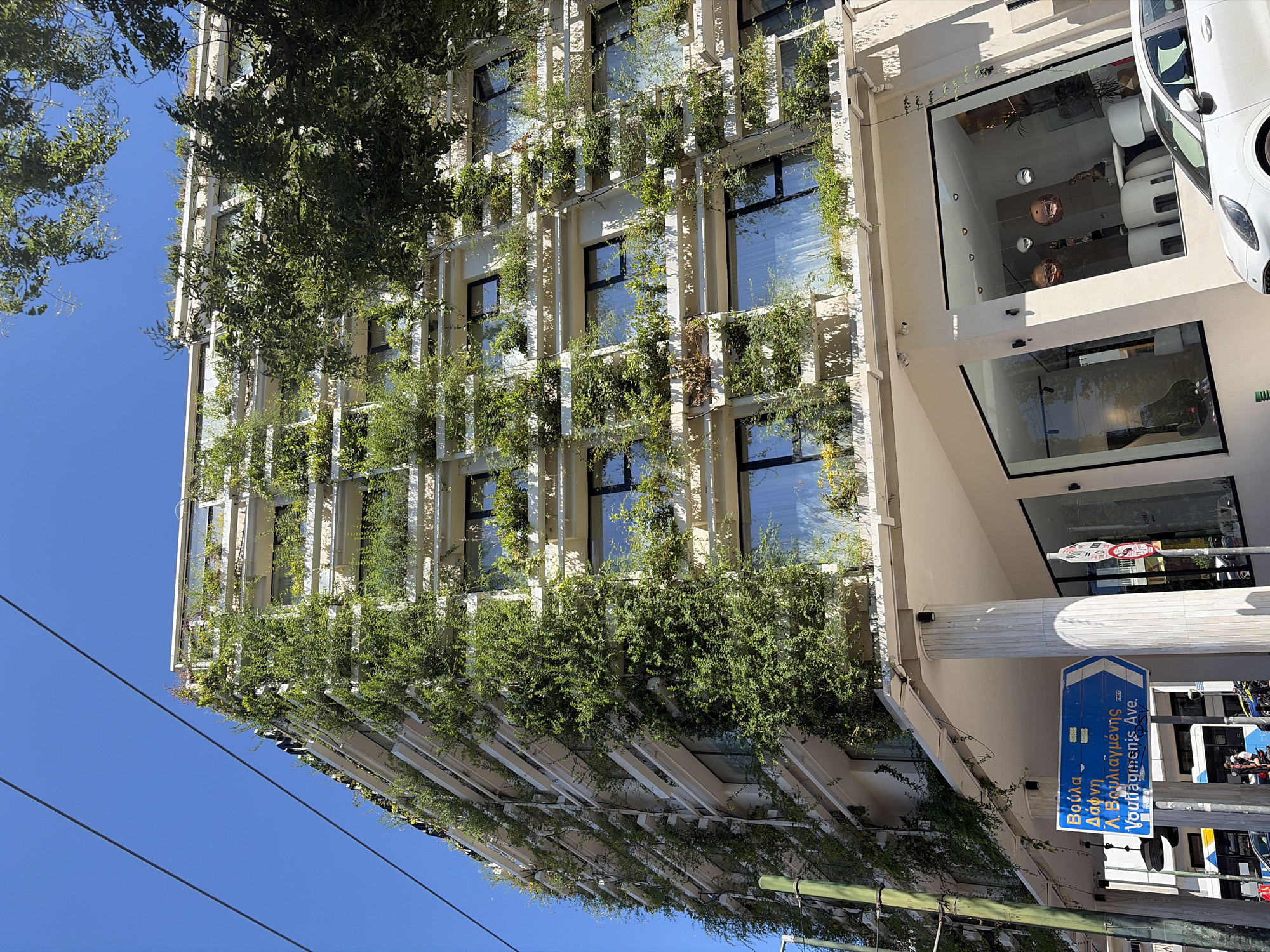 The image shows a modern building with a facade covered in lush green vines, creating a striking vertical garden effect. The building has a contemporary architectural design with numerous windows and light-colored structural elements. Sunlight illuminates the scene, suggesting it is daytime with clear weather.
On the right side of the building, a white car is partially visible, parked alongside the street. Below the building, a street sign with blue and white markings is prominently displayed. The sign contains text in both Greek and English. The Greek text reads "Βούλα" and "Λ. Βουλιαγμένης", while the English text says "Voula" and "L. Vouliagmenis Ave." This indicates the location is likely on Vouliagmenis Avenue in Voula, a coastal suburb of Athens, Greece.
The building appears to be a commercial establishment or office space, with large glass windows on the lower floors revealing interior spaces that seem to be furnished with modern decor, including what looks like seating and decorative items. The overall impression is one of an urban setting where nature and modern architecture are integrated. The perspective of the shot is from a low angle, looking upwards towards the building. Thin cables, possibly from overhead power lines or telecommunications, are visible in the lower part of the image, crossing against the clear blue sky. No people are clearly discernible in the image.