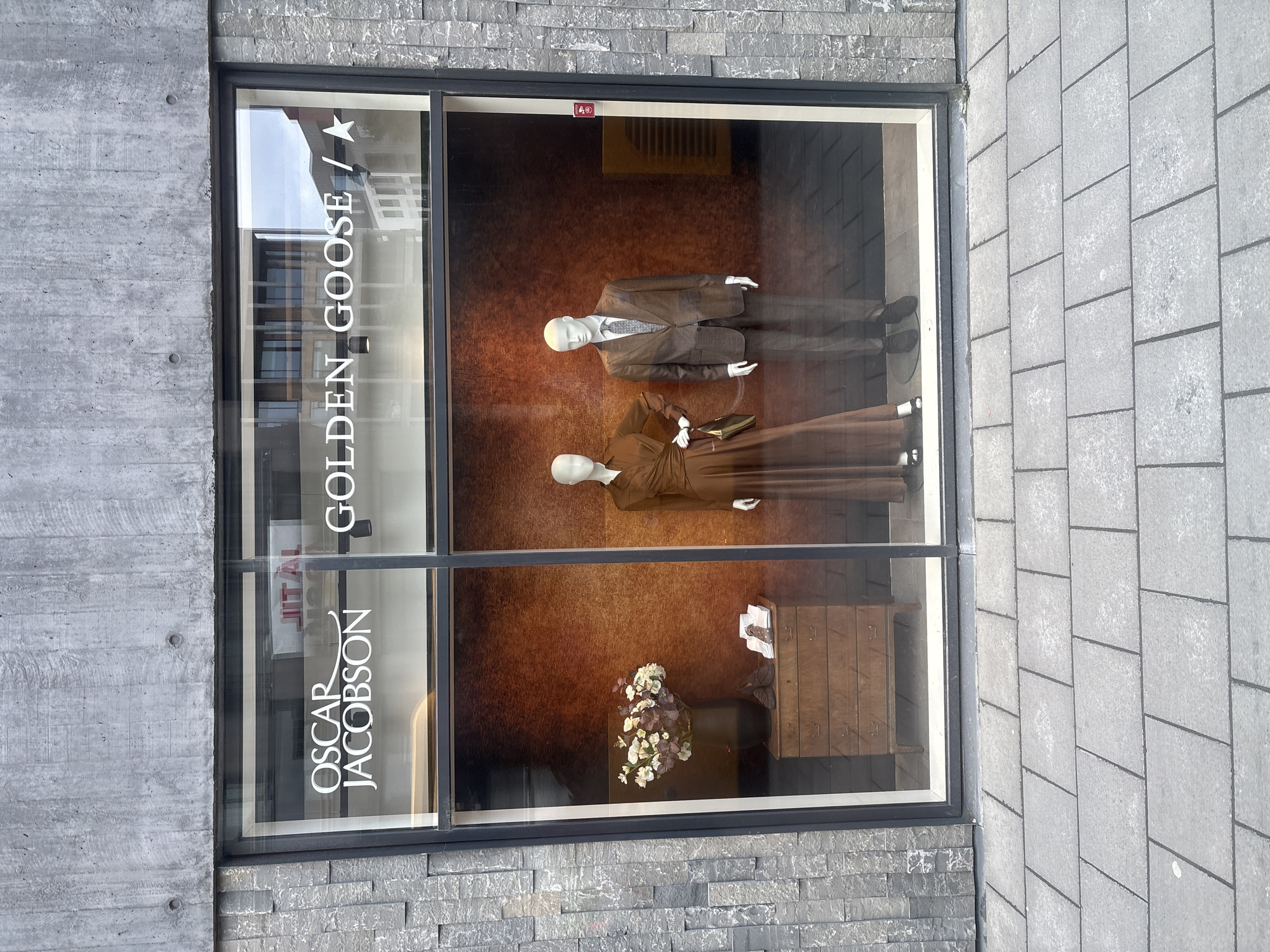 The image shows a shop window display for "OSCAR JACOBSON" and "GOLDEN GOOSE" in Reykjavik, Iceland. The window is divided into two sections. The left section displays a vase of flowers on a wooden chest of drawers, with a pair of shoes and some folded garments. The right section features two mannequins, one dressed in a long brown dress and the other in a brown suit. The storefront is constructed with grey textured stone walls and large glass windows. The pavement in front of the shop is made of grey rectangular tiles.