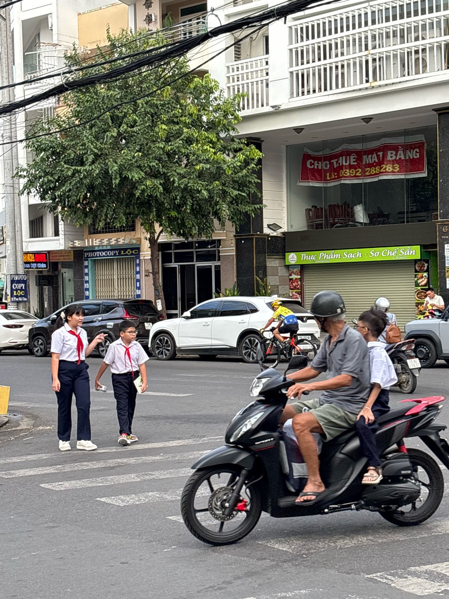 In Nha Trang, Vietnam, on a street, two young students in school uniforms with red ties are crossing a crosswalk. The female student on the left, wearing a white shirt and dark pants, is looking and gesturing to the right. The male student on the right, also in a white shirt and dark pants, is wearing glasses and holding a book. Behind them, a man and a child are riding on a black scooter. The man, wearing a gray t-shirt and shorts, is driving, while the child, wearing a white shirt and dark pants, is seated behind him. The child is wearing sandals. The environment includes buildings with signs, trees, and other vehicles such as cars and a bicycle in the background. The scene appears to be during daylight.