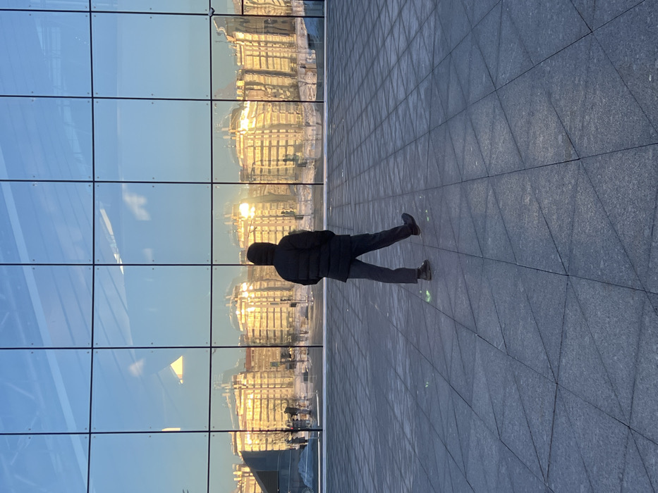 An outdoor wide shot in Saint Petersburg, Russia, depicts a single individual walking away from the viewer towards a modern building with a expansive glass facade. The person wears a dark, hooded puffer jacket, dark trousers, and dark shoes, with their right foot raised mid-stride. They are situated on a large, dark gray paved plaza featuring a geometric pattern of stone tiles, some of which exhibit small, bright green light glints near the individual's feet.
The towering glass facade, composed of a grid of square and rectangular panels, dominates the background, acting as a reflective surface. It prominently displays the reflection of multiple multi-story classical-style buildings, uniformly illuminated by a strong golden light, indicative of either sunrise or sunset. Snow is visible on the ground in front of these reflected buildings, confirming a winter season. The upper portion of the reflection shows a clear blue sky. The scene captures a solitary figure in a crisp, clear urban environment, juxtaposing contemporary architecture with the reflected historic cityscape characteristic of Saint Petersburg.