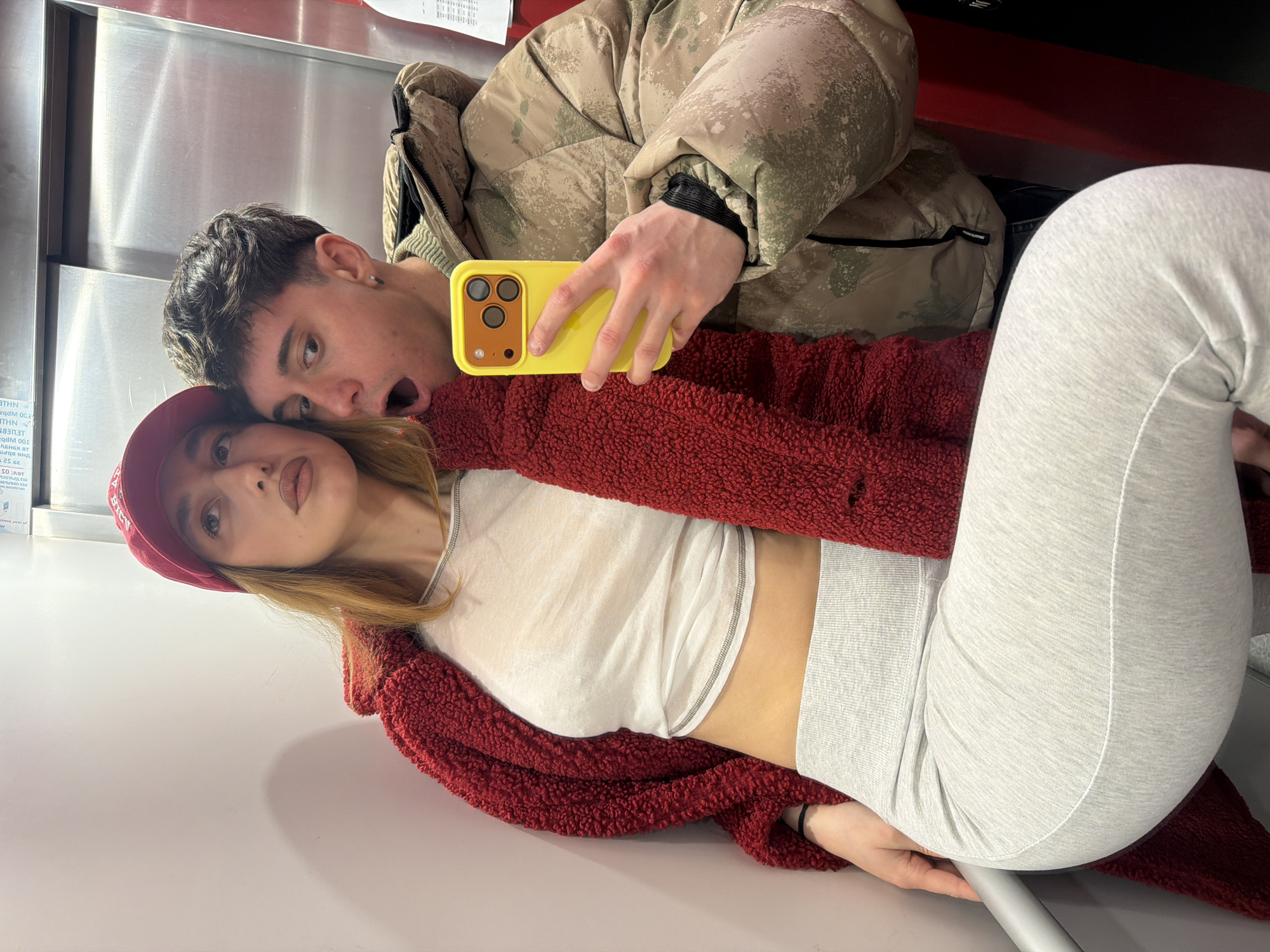 A rotated eye-level shot captures a couple taking a selfie inside what appears to be a modern elevator. The woman, wearing a red cap, a white top, gray leggings, and a red cardigan, is looking at the camera. The man, sporting a camouflage jacket, is holding a yellow iPhone and making a surprised expression with his mouth open wide. The environment is minimalistic, featuring metallic walls and modern surfaces. In the background, there's a sticker with "WHN" and other text. Based on limited information, the location cannot be definitively determined as Sofia, Bulgaria, without additional context or identifying landmarks.