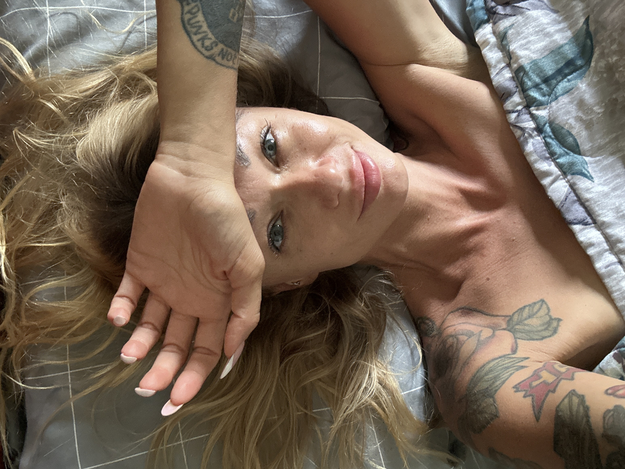 A woman with blonde hair and blue eyes is lying in bed, her hand covering her forehead. She is wearing a grey and white patterned comforter. Tattoos are visible on her left arm and shoulder. The bed is covered in a grey, grid-patterned sheet. The scene appears to be indoors.