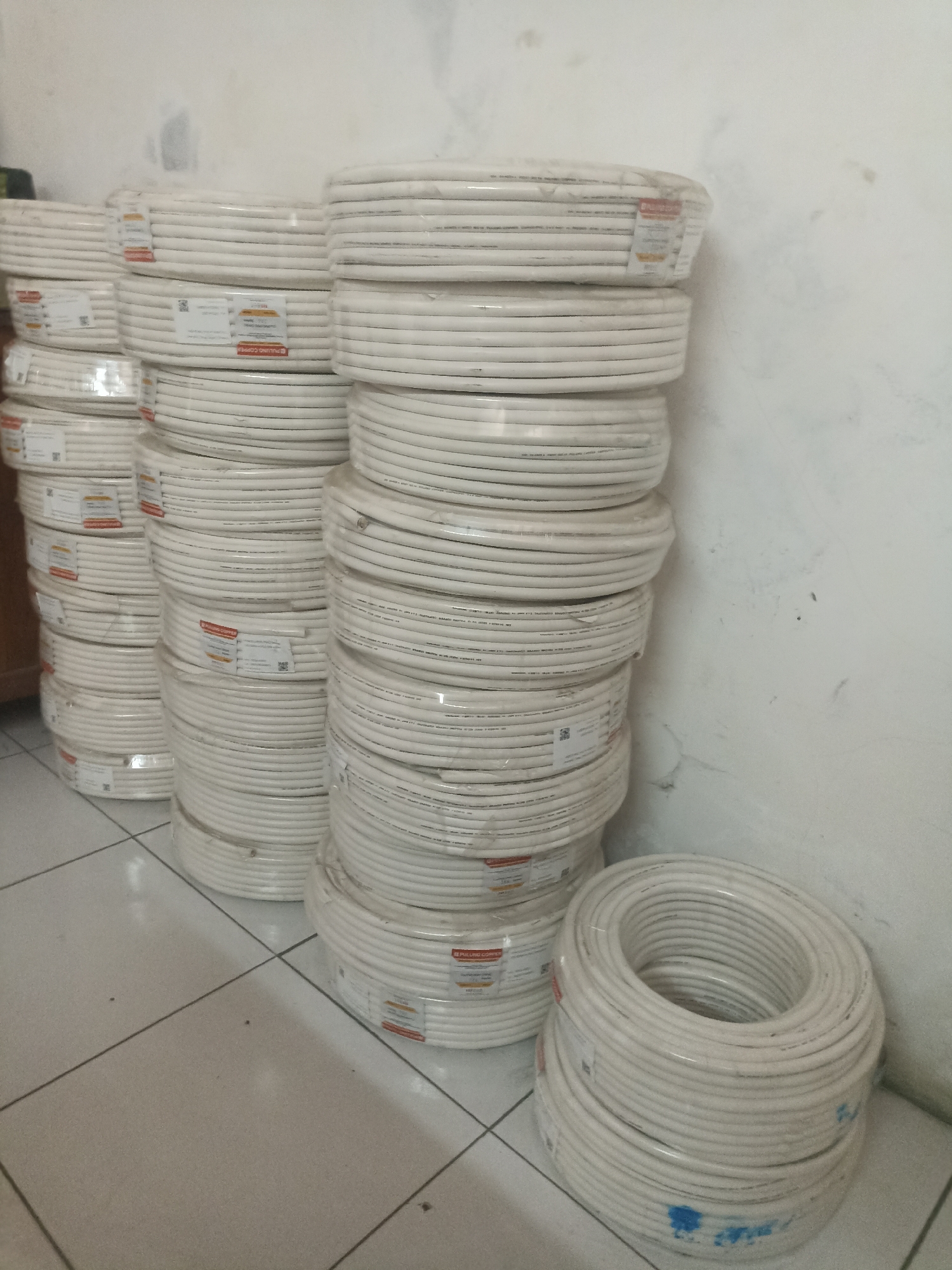 The image depicts a storage area in Surakarta, Indonesia, featuring stacks of coiled white cables, likely electrical or coaxial cables, wrapped in clear plastic. The cables are arranged in multiple vertical columns, with some coils also placed horizontally on the floor. Labels are visible on some of the cable rolls, though the text is largely illegible due to the image resolution. The environment appears to be an indoor space with tiled flooring and a plain white wall in the background. The lighting is somewhat dim, casting shadows. There are no people or animals present in the scene. The overall impression is one of a stock of goods awaiting distribution or sale.