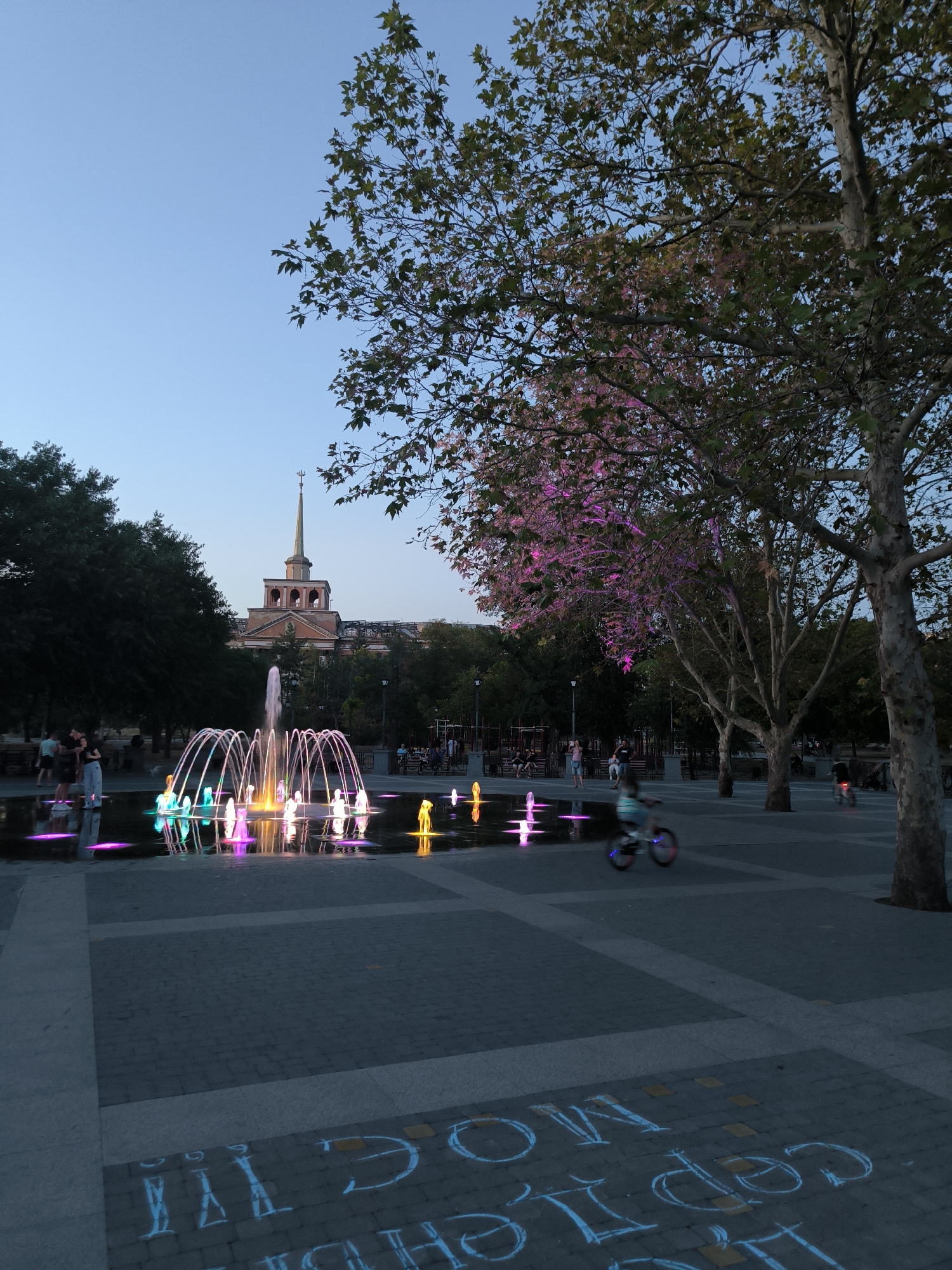 The image shows a public square in Mykolaiv, Ukraine, featuring a modern fountain with colorful LED lights. The fountain is the central element, with streams of water illuminated in various hues of pink, purple, green, and yellow, reflecting on the dark surface of the water basin. In the background, a prominent building with a distinctive spire rises above the trees, characteristic of Mykolaiv's architectural style.
Several people are visible in the scene. On the left, a couple stands near the fountain. Further back, a child rides a bicycle with blurred motion, suggesting movement. Other individuals are seated on benches or walking in the background, indicating a leisurely evening atmosphere.
The square is paved with dark cobblestones, with lighter colored square sections creating a patterned walkway. Some of the paving stones have chalk drawings on them, including what appear to be Cyrillic letters.
Lush green trees surround the square, with one large tree in the foreground on the right, its branches partially obscuring the view. The tree on the left side of the image is also large and leafy, with some branches illuminated by pink light, possibly from a nearby source or the fountain itself. The sky above is a clear, deep blue, indicating either early evening or late twilight. The overall scene conveys a sense of a well-maintained public space designed for recreation and relaxation.