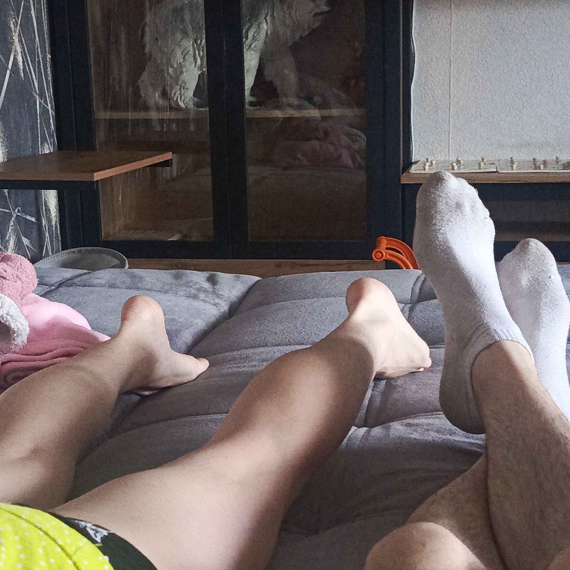 The image shows two pairs of legs resting on a gray quilted surface. One pair is bare-footed while the other is wearing white socks. The bare-footed legs are on the left and belong to someone wearing yellow shorts with white polka dots. Behind them is a cabinet with glass doors, a wooden shelf, and a curtain. A toy is visible behind the socked feet. The chess board is on a wooden shelf on the right side of the frame. The location in the city of Марковский, Russia is assumed.