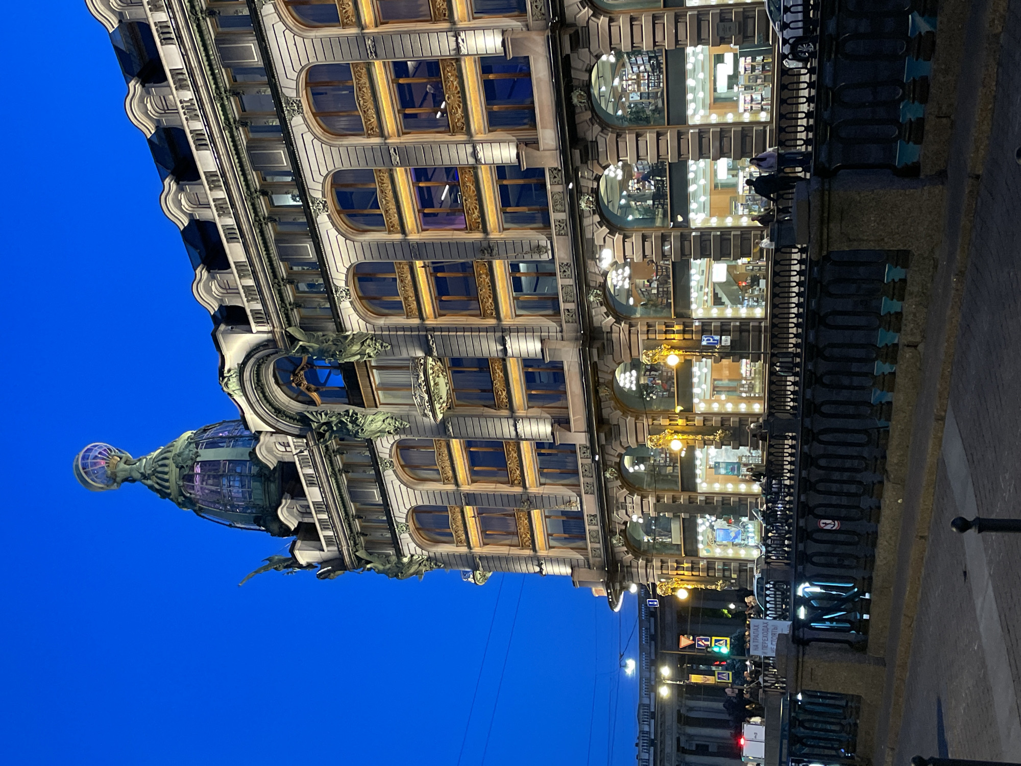 The image displays an ornate, multi-story building at dusk or night, illuminated by its interior lights and streetlamps, set against a deep blue sky. The building exhibits a rich architectural style, featuring an elaborately decorated facade with intricate carvings, including gargoyle-like figures. Its upper floors have numerous windows, some arched and some rectangular, with golden decorative panels between them. A prominent feature is a large, glass dome topped with a globe-like structure, visible on the left side of the building.
The lower levels of the building consist of brightly lit display windows, revealing the interiors of what appears to be a department store or shops. In the foreground, a dark, decorative metal railing runs alongside a paved area, suggesting a street or embankment next to a canal or river. Golden-hued streetlights dot the scene, casting light onto the pavement. Several blurry figures, presumably people, are visible near the railing and on the street, some standing or walking. Faint power lines crisscross the twilight sky. A white banner attached to the railing displays Russian text: "НА ТРАПАХ ПЕРЕХОДАХ НЕ СТОЯТЬ," which translates to "DO NOT STAND ON GANGWAYS/TRANSITIONS."