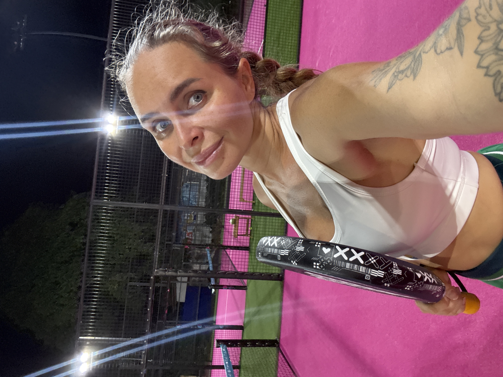 A woman with blonde hair in a braid, wearing a white sports bra, is taking a selfie on a brightly lit, pink padel court at night. She is holding a black and white padel racket. The court has green borders and is surrounded by a fence and stadium lights. The location is Ko Samui District, Thailand.
