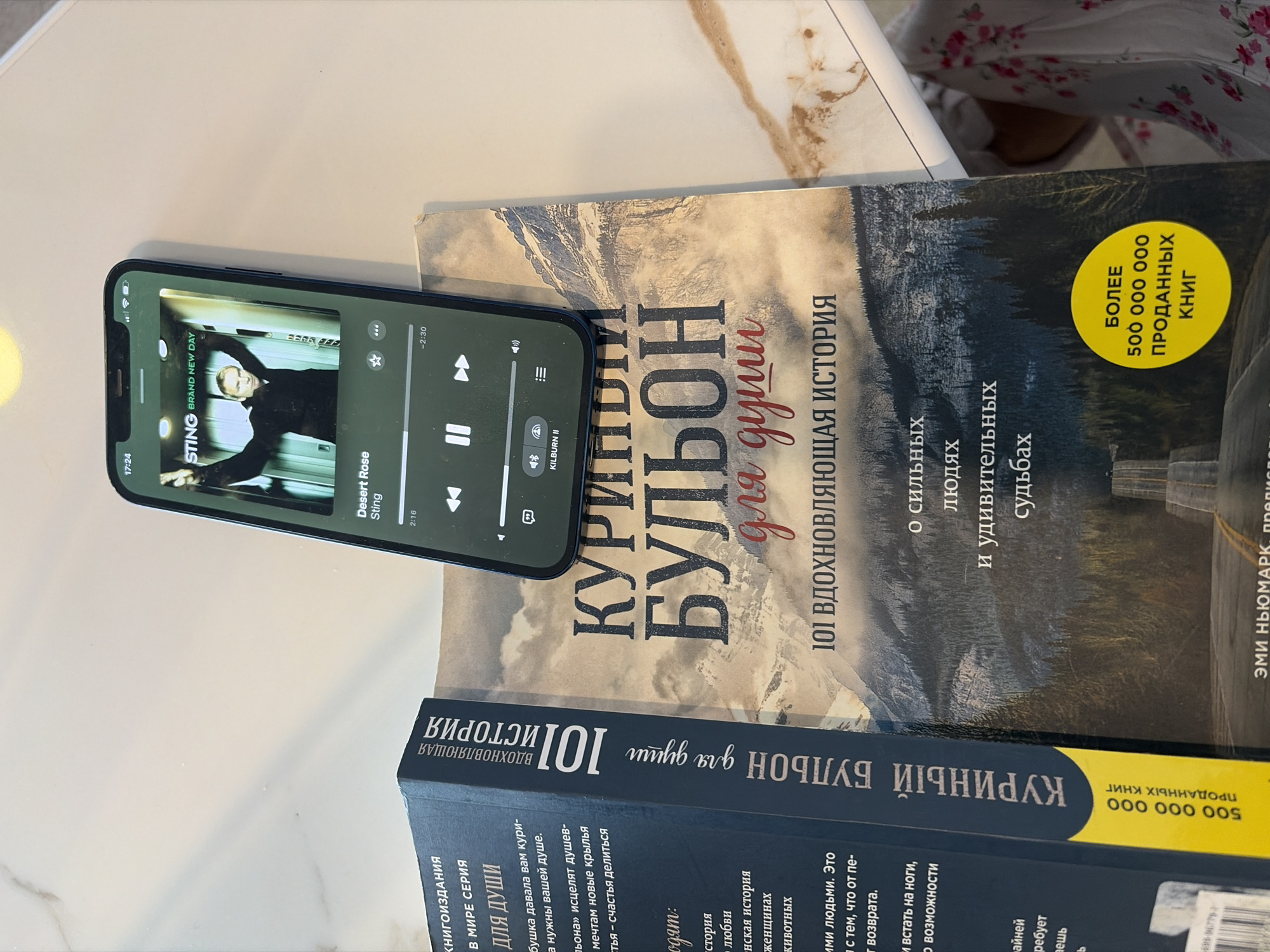 A mobile phone is placed on top of two stacked books. The phone screen displays a music player application, showing the song "Desert Rose" by Sting. The top book, "Chicken Soup for the Soul," has a mountain landscape cover, with the title in Cyrillic. The book states "101 Inspirational stories" and "more than 500 million books sold". The bottom book is the same title but only the spine is visible, in Cyrillic. The books and phone are on a white table, possibly made of marble, with a glimpse of fabric with a floral pattern.