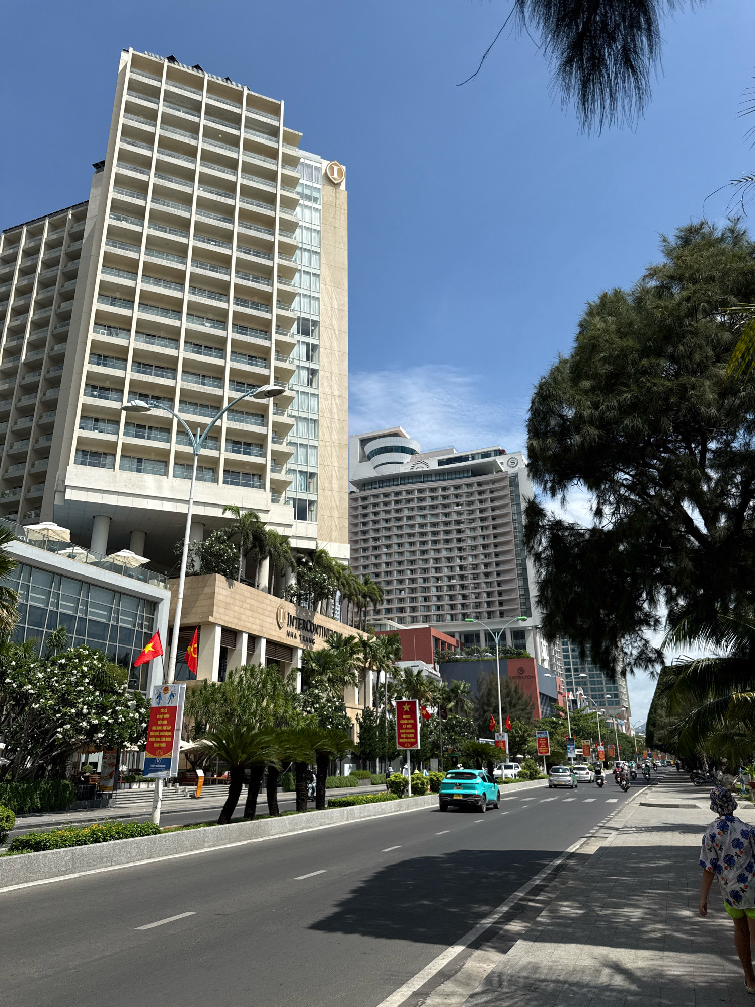 The image depicts a street scene in Nha Trang, Vietnam, featuring several high-rise buildings. Dominating the left side of the frame is a tall, cream-colored hotel with numerous balconies and a large "I" logo on its upper facade. Adjacent to it is another, wider hotel building with many windows. Further down the street, a more modern, grey building with a distinctive curved roofline is visible. A wide, paved road runs through the center of the image, with traffic including cars, motorcycles, and a turquoise car. On the right side of the road, a person wearing a hat and a patterned shirt walks on the sidewalk. Trees and palm trees line the street, and flags, including Vietnamese flags, are displayed. The sky is clear blue with some wispy clouds. The scene suggests a sunny day in a coastal city.