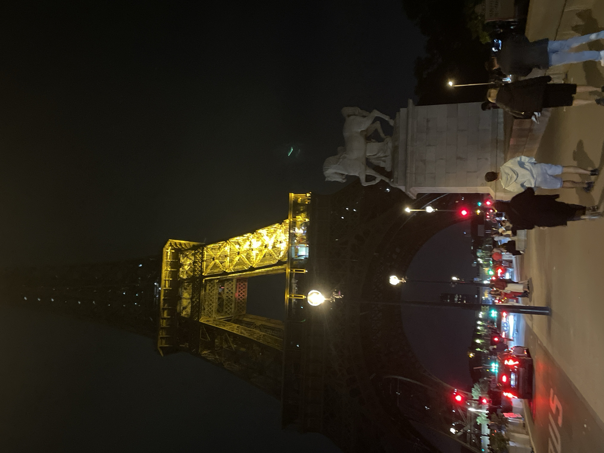 The image captures a nighttime scene in Paris, France, featuring the Eiffel Tower prominently illuminated in the background. The Eiffel Tower is a wrought-iron lattice tower located on the Champ de Mars in Paris. In the foreground, a street with traffic lights is visible, with cars driving by. Several people are walking along a pathway. To the right of the Eiffel Tower, a large stone sculpture of a horse stands on a pedestal. The overall atmosphere is that of a busy urban environment at night, with artificial lighting from the tower, streetlights, and vehicles illuminating the scene against a dark sky.