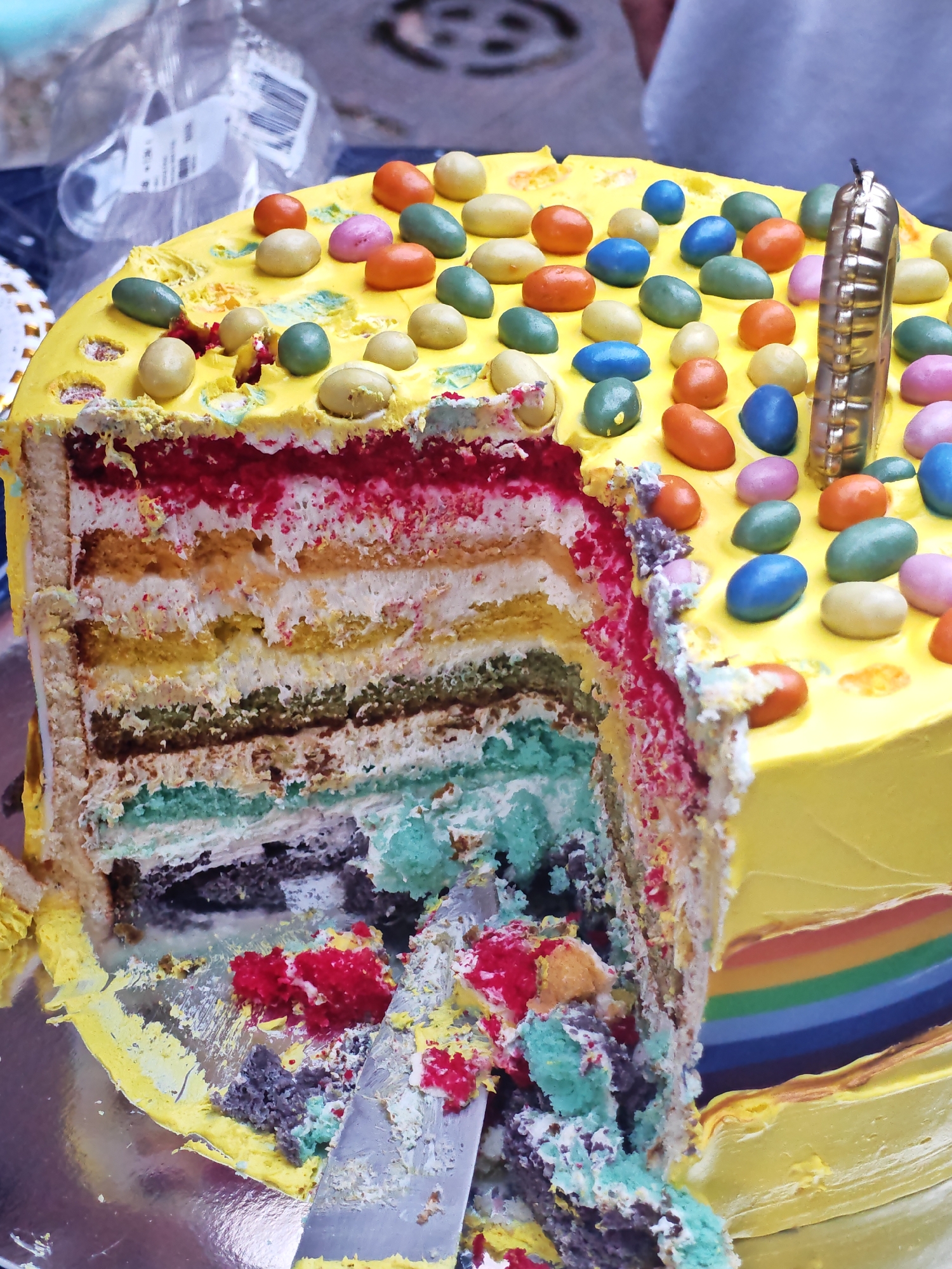 A sliced, multi-layered rainbow cake is displayed on a metallic surface. The cake is frosted in bright yellow, with a rainbow stripe visible on its side. The top of the cake is adorned with colorful candy-coated chocolates and a single gold candle. A slice has been removed, revealing the cake's vibrant interior layers of red, orange, yellow, green, and blue. Crumbs and frosting are scattered around the removed slice. The background is blurred, suggesting an outdoor setting. There is no visible indication of a specific location in Sofia, Bulgaria.