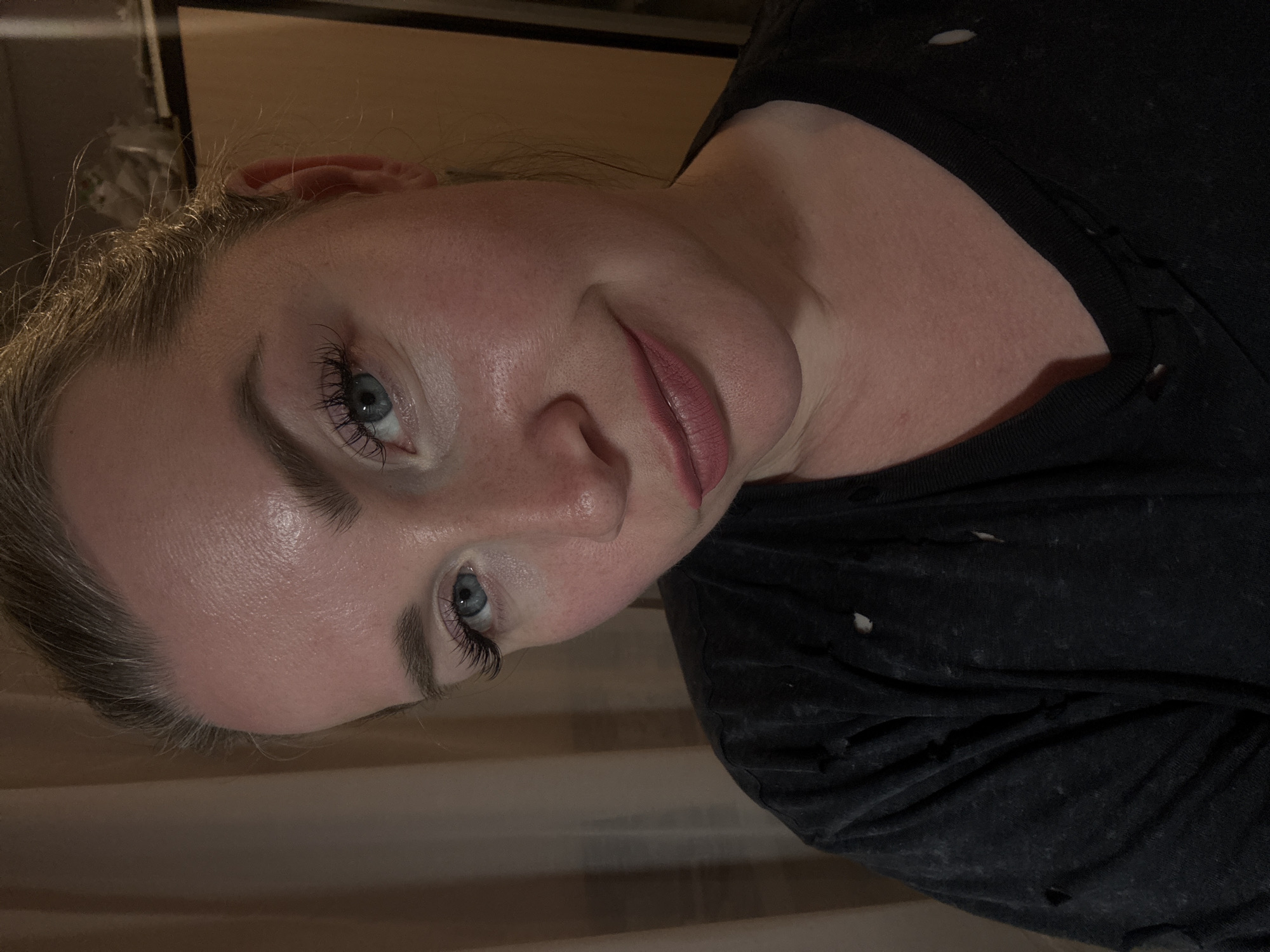 A medium close-up, chest-up portrait shows an adult female indoors. She has fair skin with visible pores and some redness, particularly on her cheeks and forehead, which also exhibits a slight sheen. Her eyes are blue with prominent, long dark eyelashes, and her eyebrows are light brown and well-defined. Her light-colored hair is pulled back, with some wisps visible at the hairline. Her lips are full and colored with a matte reddish-pink lipstick, forming a subtle, closed-mouth smirk. She is wearing a dark, possibly charcoal or black, V-neck top that appears distressed with small holes and frayed edges. The background to the left consists of soft, light-brown draped fabric, likely curtains. To the right, the background is darker and indistinct, with a vertical element suggesting a piece of furniture or a door frame, and some blurry, lighter objects resting on a surface in the upper right corner.