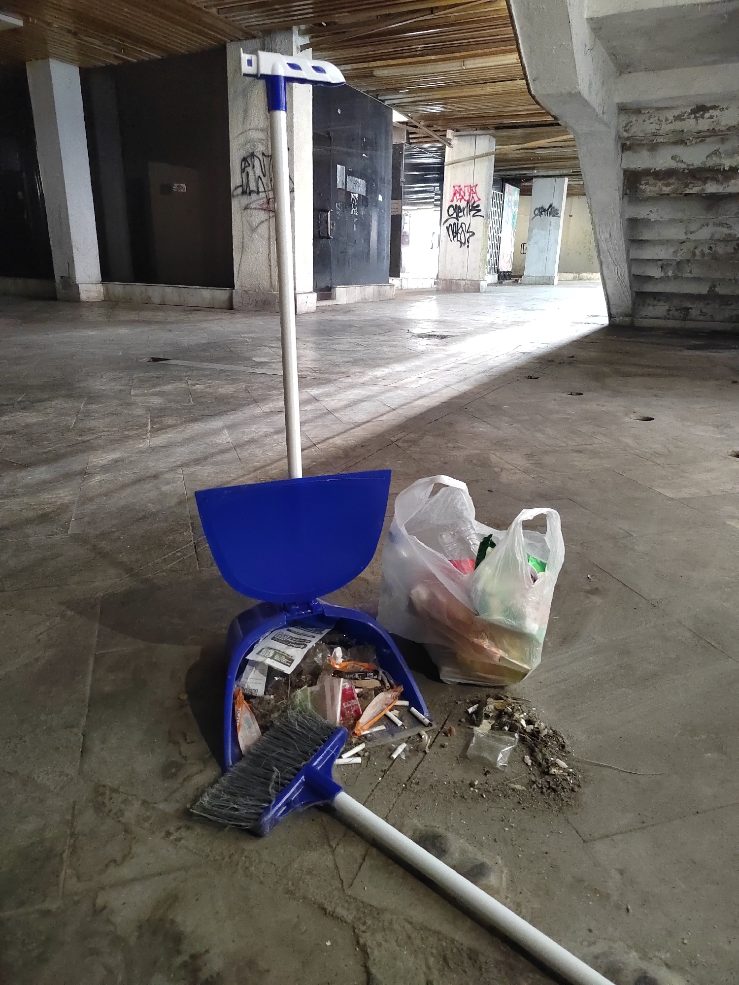 The image depicts a cleaning setup in a large, concrete space in Podgorica, Montenegro. The space appears to be an unfinished or abandoned area, with exposed wooden beams overhead, concrete pillars, and a concrete floor. Graffiti is visible on the pillars. A blue dustpan is holding trash, including cigarette butts and small wrappers. A broom with a blue head and white handle is on the floor next to the dustpan. A clear plastic bag filled with trash sits behind the dustpan. The scene suggests ongoing or recently completed cleaning in a neglected area. The lighting is dim, with natural light filtering in from the open space.