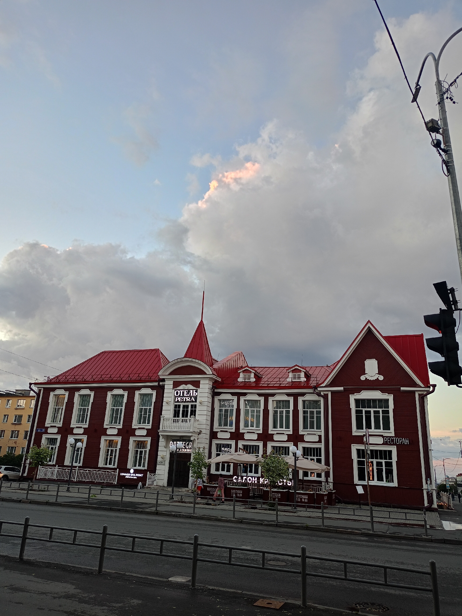The image shows a three-story red wooden building with white trim and a red roof. The building has a prominent central section with a white facade and a pointed spire. Signs on the building indicate "ОТЕЛЬ PETRA" (Hotel PETRA) and "РЕСТОРАН" (Restaurant). There are several windows on each floor, many with white frames. To the left of the main building, a portion of a yellow apartment building is visible. In front of the building, there is a sidewalk with several people walking, and a street with a traffic light. The sky is overcast with dramatic clouds, and the lighting suggests it is late afternoon or early evening.