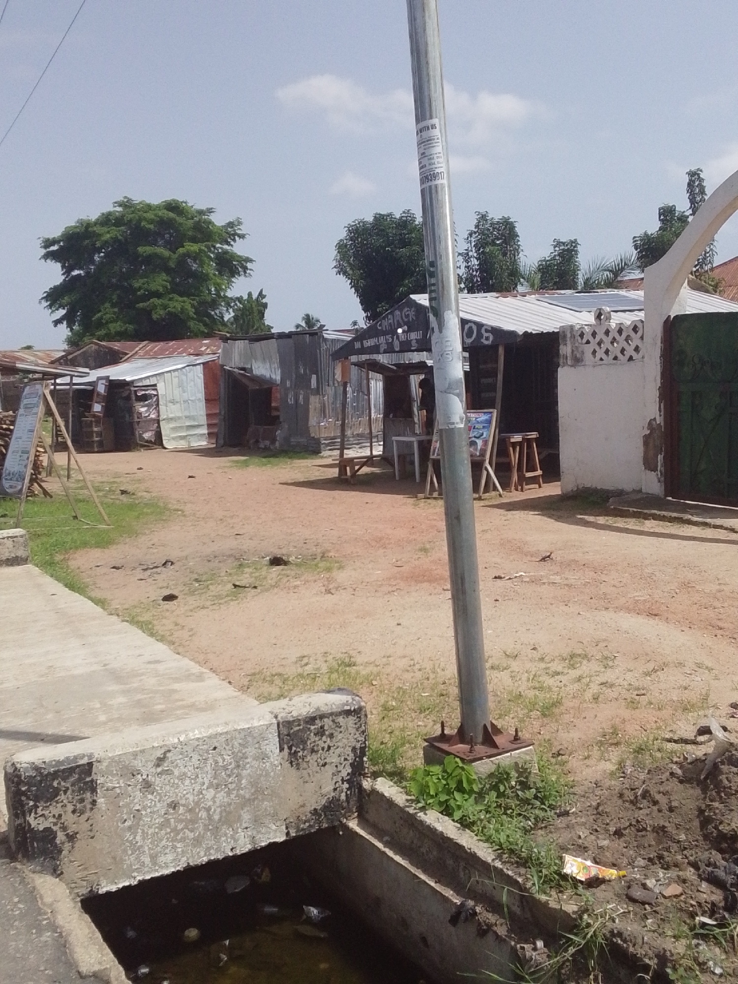 The image depicts an outdoor scene in Jalingo, Nigeria, under a clear, bright sky suggesting daytime. The setting appears to be a commercial area with several small, makeshift shops constructed from corrugated metal and wood. These structures are arranged along a dirt road, with some exhibiting signs of age and wear. Solar panels are visible on the roof of one of the buildings, indicating a modern addition to the otherwise rustic environment.
In the foreground, a concrete structure, possibly part of a drainage system, is prominent. The surrounding ground is a mix of dirt and sparse green vegetation. Several objects are scattered around, including what looks like discarded materials and a small lizard on the ground. A tall metal pole, possibly a lamppost or utility pole, stands near the center of the image, adorned with several stickers or flyers. One sticker displays text, including what appears to be a phone number "0807939017" and "WITH US". Another sign or poster on an easel is visible to the left, and a sign above one of the shops reads "CHARGE" and "OS".
The overall impression is one of a bustling, informal marketplace or commercial strip in a developing urban or semi-urban area. The architecture and the arrangement of the shops suggest a local, community-oriented business environment. The clear sky and bright sunlight indicate fair weather. No people are clearly visible in the image, but the presence of the shops and signage implies ongoing commercial activity.