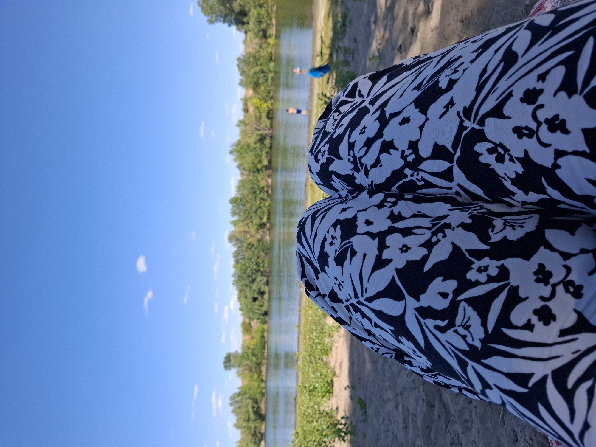 The image depicts a close-up view of a person's legs, wearing black and white floral patterned pants, resting on a sandy shore. In the background, a wide river is visible, with a line of green trees and vegetation along its banks. The sky is clear blue with a few scattered white clouds. Two small figures, seemingly people, are in the river, one in blue shorts and the other in darker attire. A blue object, possibly a bag, is on the sandy bank near the river. The scene suggests a relaxed atmosphere by the water on a sunny day. The location is identified as Dubăsarii Vechi, Moldova.