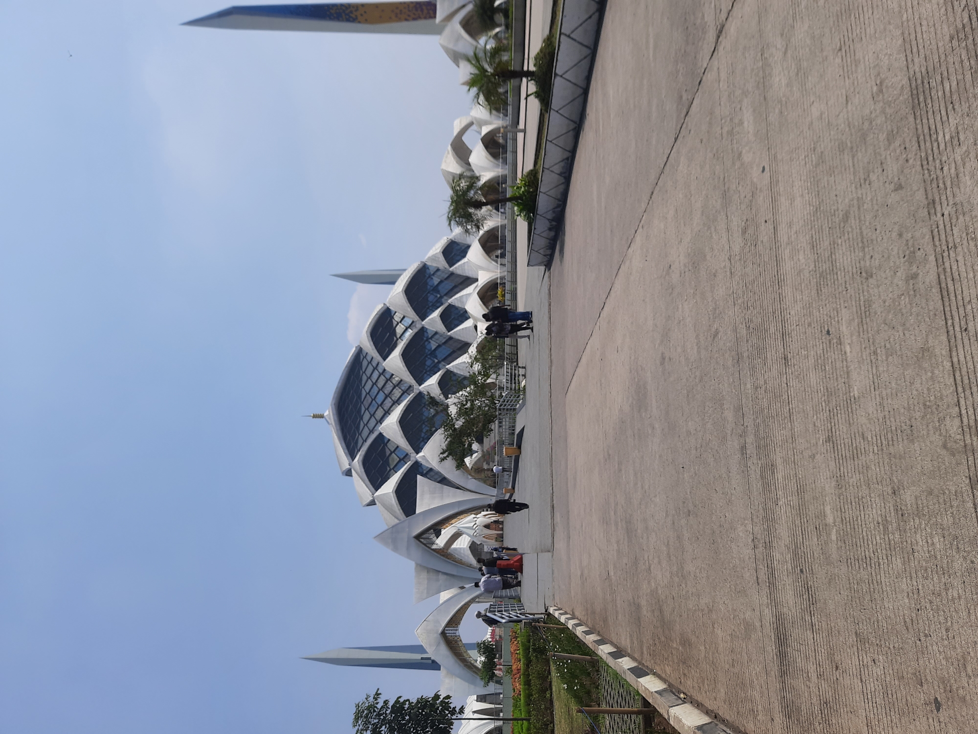 This image shows the exterior of the Gelora Bandung Lautan Api Stadium in Bandung, Indonesia, captured from a slightly elevated position looking towards the main entrance. The sky is clear and blue, with a bright, sunny day indicated. In the foreground, a concrete pathway with visible tire marks leads towards the stadium. On either side of the pathway, there are low concrete borders with black and white striped edging.
The Gelora Bandung Lautan Api Stadium itself is a prominent structure, characterized by its unique, modern architectural design. The stadium features a large, curved roof with a diamond-shaped pattern, supported by numerous white, petal-like structures that arch upwards. Two tall, slender spires flank the stadium, one on the left and another on the right, with the right spire displaying a distinctive yellow and blue pattern.
Several individuals are visible in the middle ground, walking on a raised walkway leading into the stadium. Some are standing and conversing, while others are walking away from the camera. There are also palm trees and other greenery planted in planters along the walkway and in front of the stadium. The overall impression is a grand, modern stadium set against a clear blue sky, suggesting a public gathering or event.