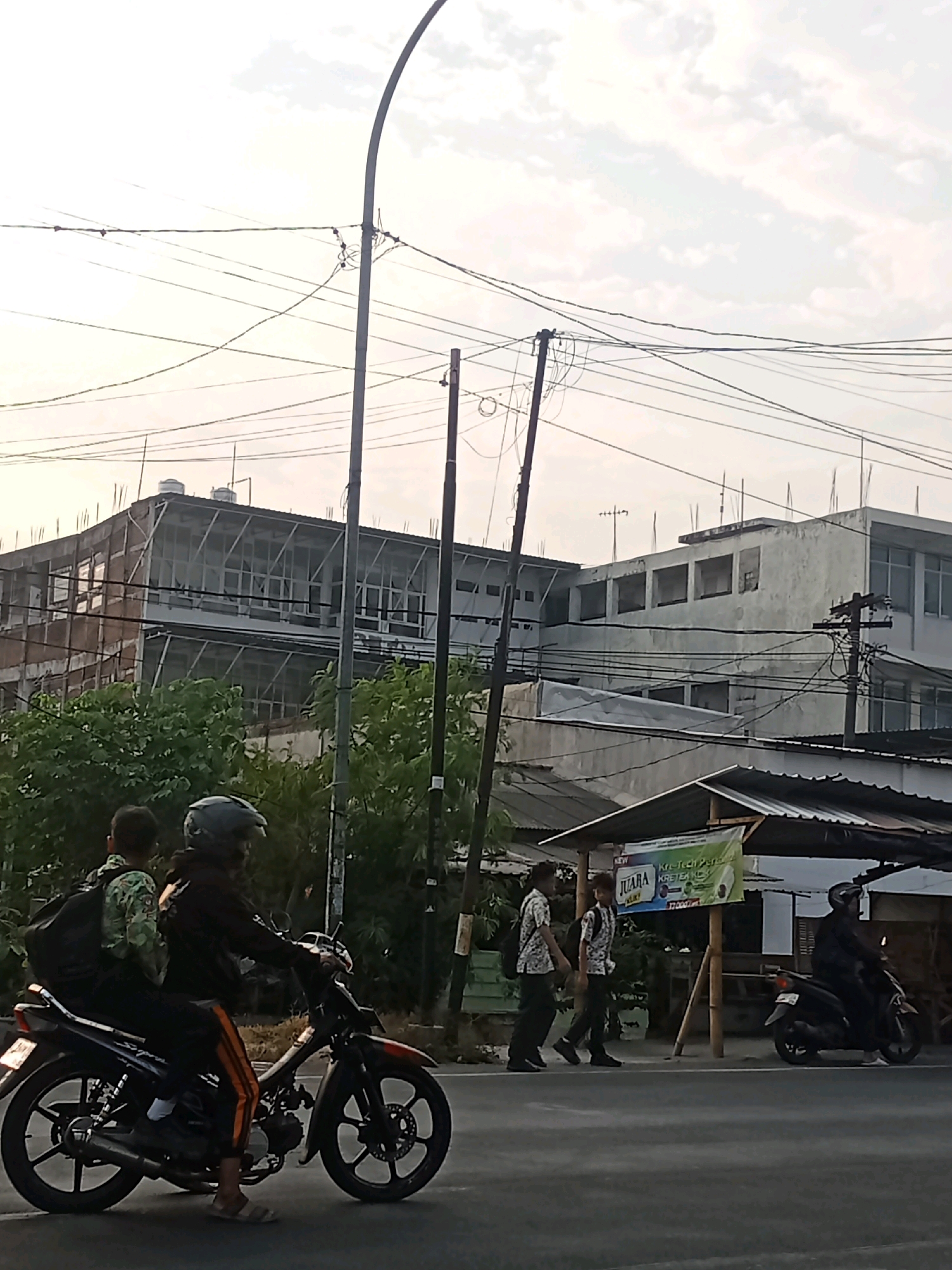 The image captures a street scene in Kaliwungu, Indonesia, under a hazy sky with scattered clouds. In the foreground, a motorcycle with two occupants, a driver wearing a helmet and a passenger with a backpack, is moving on the asphalt road. The passenger is wearing a patterned shirt. To the left, a building under construction with exposed brickwork and large glass panels is visible, surrounded by lush green trees. Across the street, a small structure with a corrugated roof serves as a roadside stall or shop, displaying a banner with text and an image. Two young men in school uniforms are walking on the sidewalk near the stall, one carrying a backpack. Further down the road, another motorcycle with a rider in a helmet is parked. Overhead, a complex network of power lines crisscrosses the sky, supported by utility poles. The overall impression is one of a bustling urban environment with elements of daily life and ongoing development.