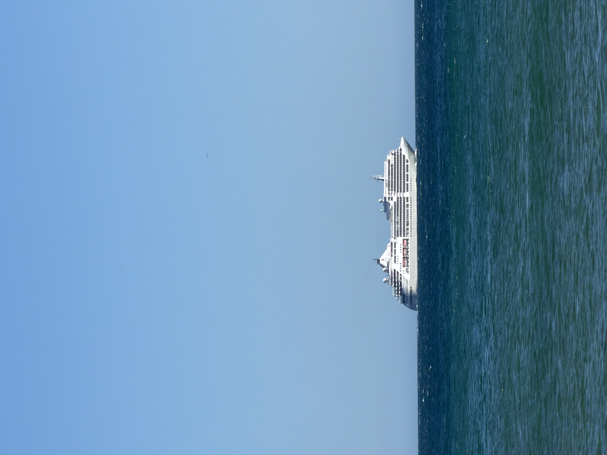 The image depicts a cruise ship sailing on the ocean off the coast of Cascais, Portugal. The ship is positioned towards the right side of the image, contrasting with the vast expanse of the clear blue sky on the left. The ocean is a dark green color.