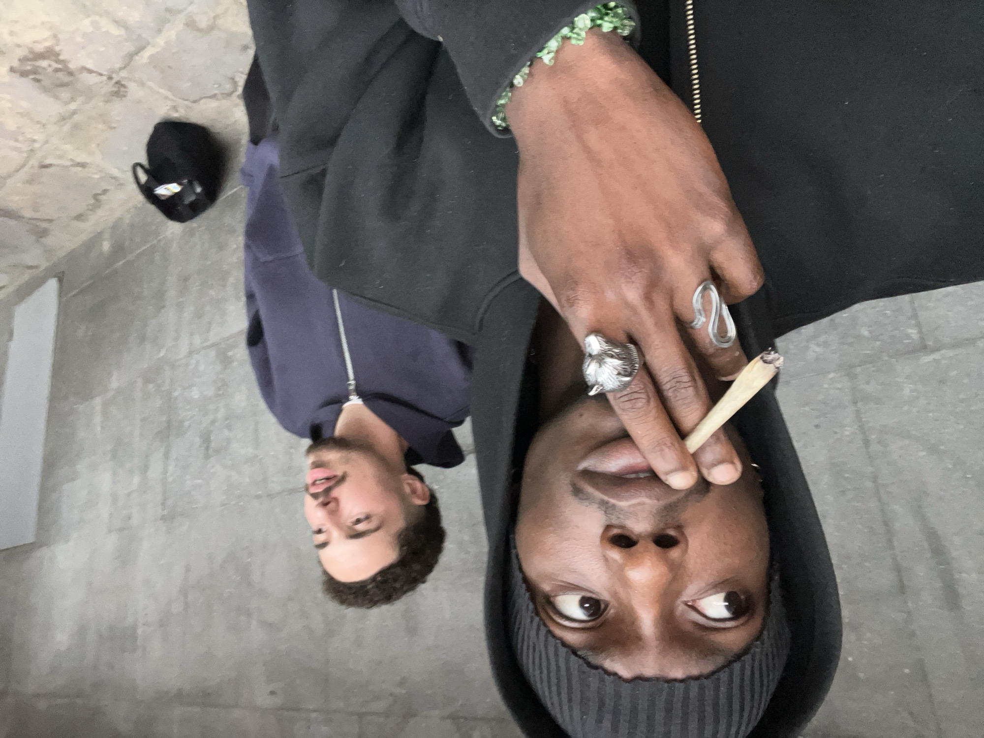 An overhead shot captures two men outdoors, seemingly lying on the ground looking up at the camera. In the foreground, a man with dark skin, wearing a black hoodie and a dark grey beanie, holds a lit hand-rolled cigarette or joint between the fingers of his right hand, close to his mouth. He has dark eyes and a relaxed, upward gaze. His left hand is visible, adorned with a green beaded bracelet around his wrist, a prominent silver wolf-head ring on his index finger, and a distinctive wavy silver ring on his middle finger.
Behind him, slightly out of focus, a second man with lighter skin, short curly dark hair, and a beard is visible. He is wearing a dark blue zipped hoodie and has a slight smile or smirk on his face, also looking towards the camera.
The setting appears to be an urban outdoor environment, likely a street or public pathway in Barcelona, Spain, with a grey concrete or paved ground surface. A black bag or backpack is visible on the ground to the far left. The lighting suggests it is daytime, with bright, natural light illuminating the scene. There is no visible text in the image. The overall mood is casual and relaxed.