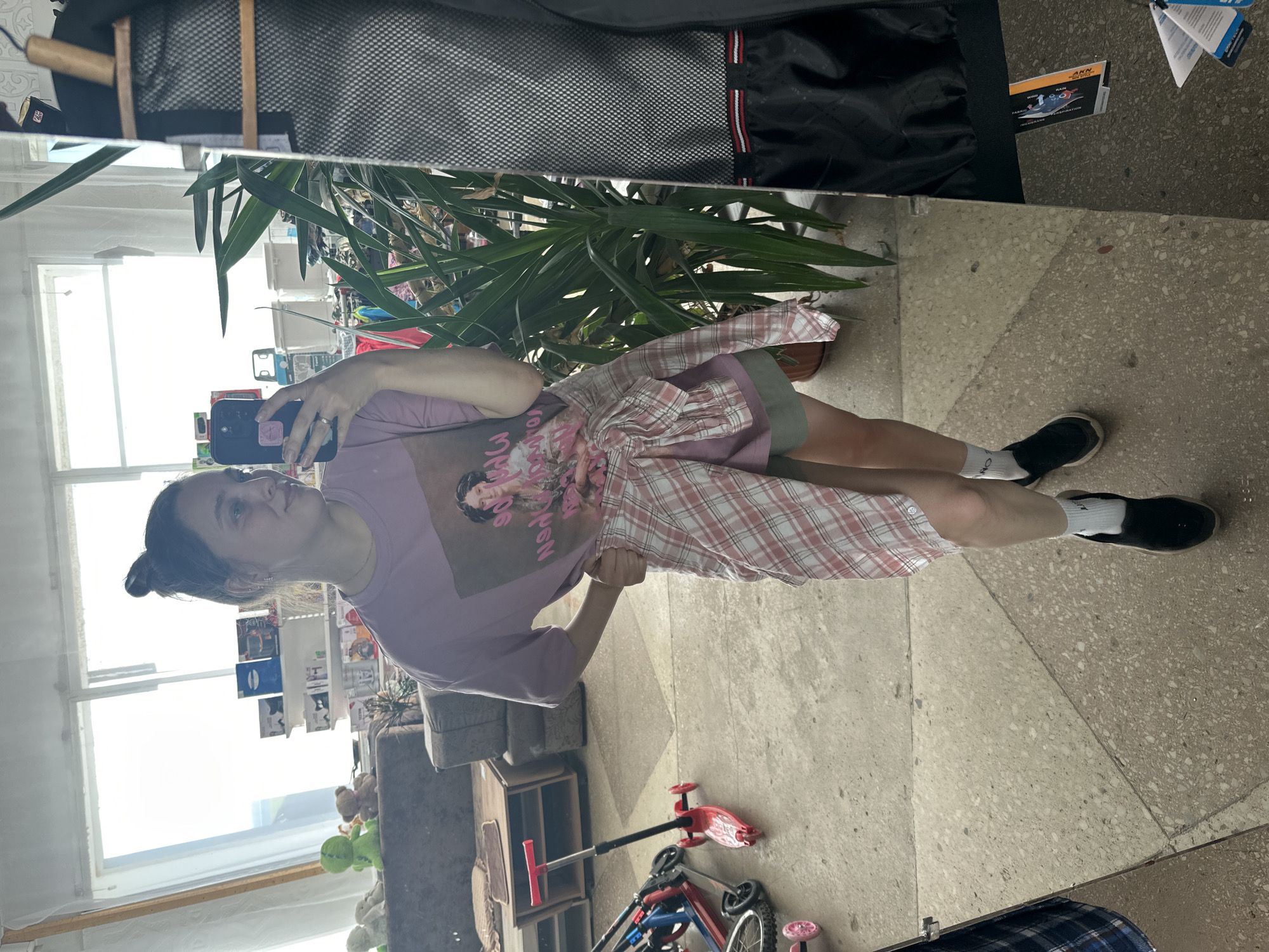 The image shows a young woman taking a mirror selfie in what appears to be an indoor space with a concrete floor, possibly a workshop or a storage area. The woman is wearing a lavender t-shirt with a graphic print, a plaid shirt tied around her waist, and green shorts. She has her hair in a bun and is wearing black slip-on shoes with white socks. In the background, there are shelves with various items, a sofa, some stuffed animals, and a large potted plant. To the left, there are several scooters, and to the right, a black jacket is hanging, with tags visible. The lighting suggests it is daytime, with light coming from a large window. The specific location of Yaroslavka, Russia, is not discernible from the visual information in the image.