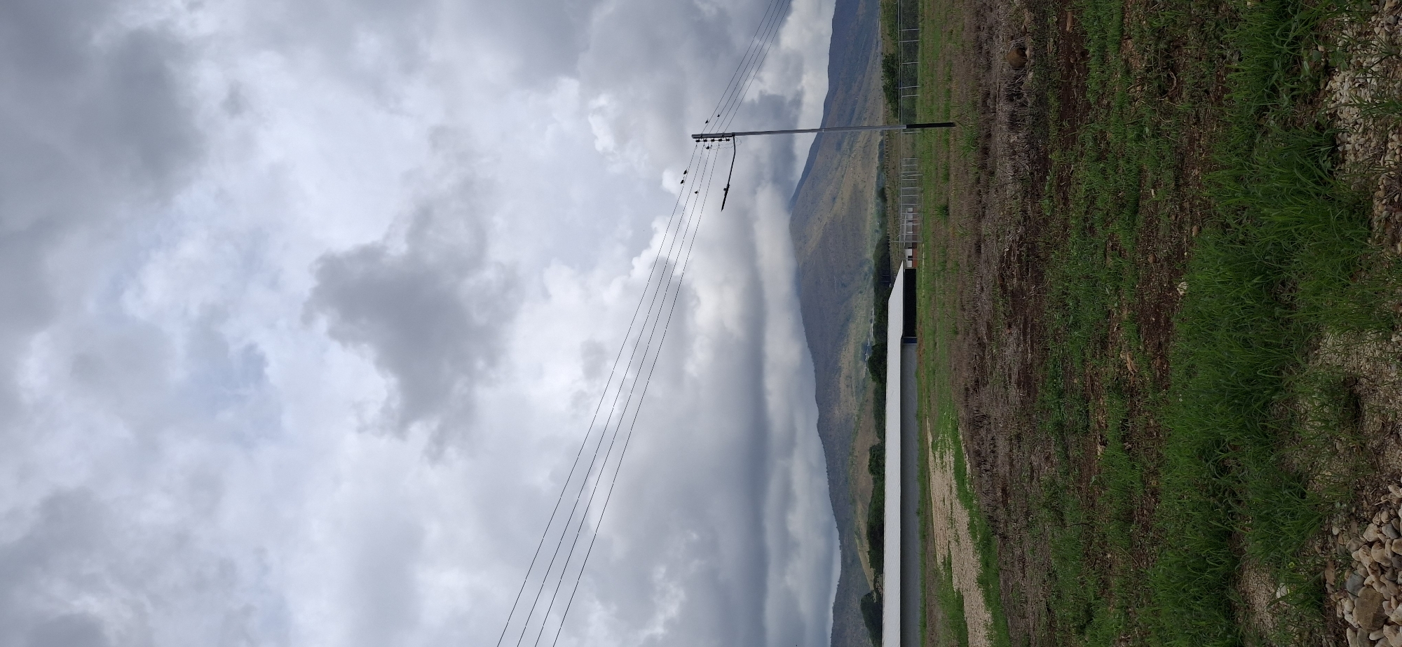 The image depicts a rural scene near Vallecito, Venezuela. The sky is overcast with grey clouds, partially obscuring the lower part of a distant mountain range. Power lines stretch diagonally across the sky. A utility pole stands upright. A long, white building runs alongside a grassy field with patches of brown earth. Pebbles are visible along the ground near the grass. A fence can be seen in the background between the building and the mountains. Several birds are perched on the power lines and pole.