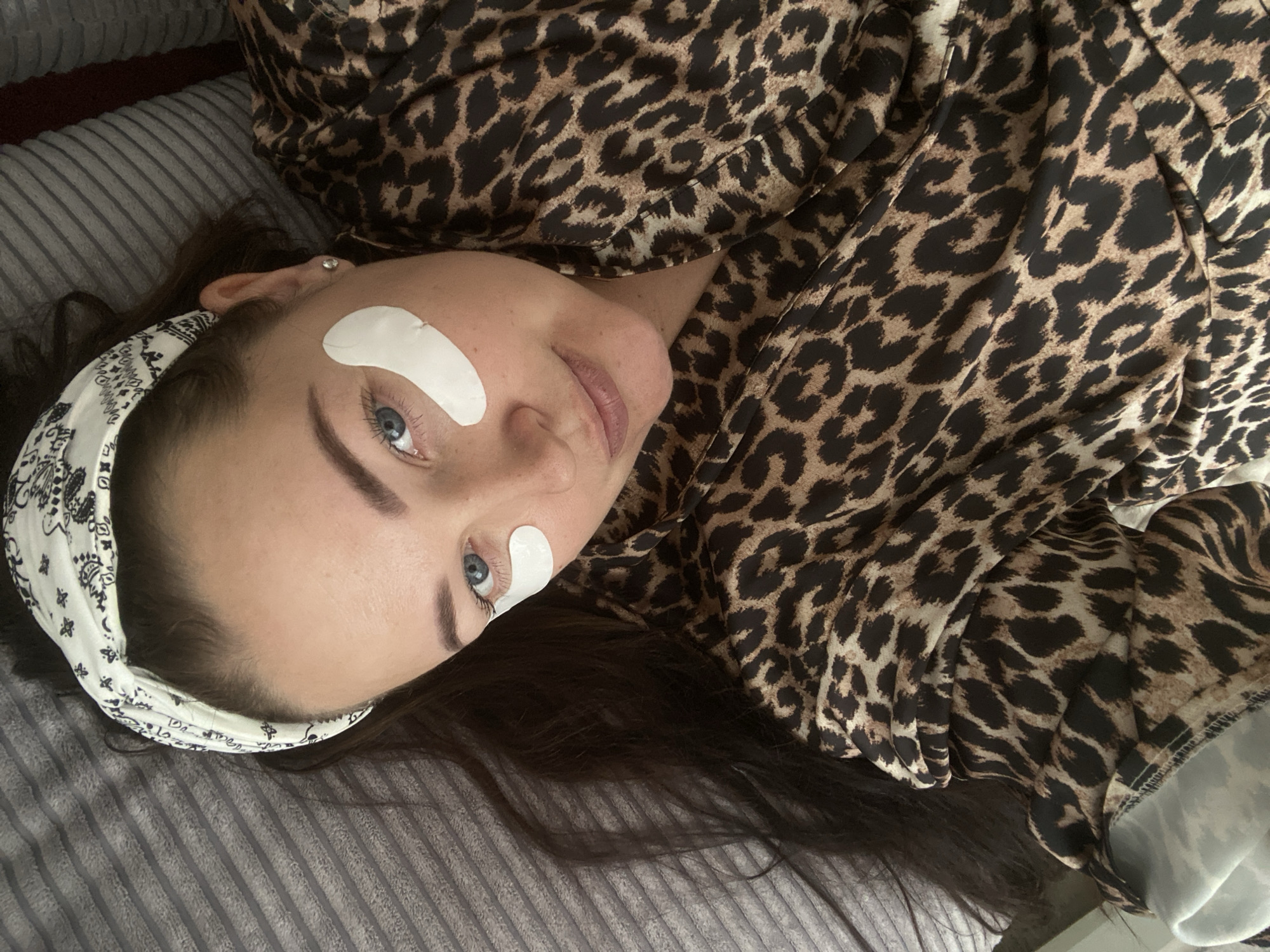 A young woman is depicted in a close-up, high-angle shot, lying on her back and looking towards the camera with a calm and soft expression. She has dark hair, striking blue eyes, and well-defined eyebrows. White, crescent-shaped patches are applied under both her eyes, suggesting a beauty or self-care treatment. She wears a white headband with a black patterned design, keeping her hair away from her face. Her attire is a prominent leopard-print top or robe, visible across a large portion of the frame. She is resting on a grey, textured blanket or bedding, which features subtle striped patterns. The scene takes place indoors, likely in a relaxed setting such as a bedroom or living area. While the image itself does not provide specific visual cues, the provided context indicates the location is Sochi, Russia. No discernible text is visible on the headband or clothing.