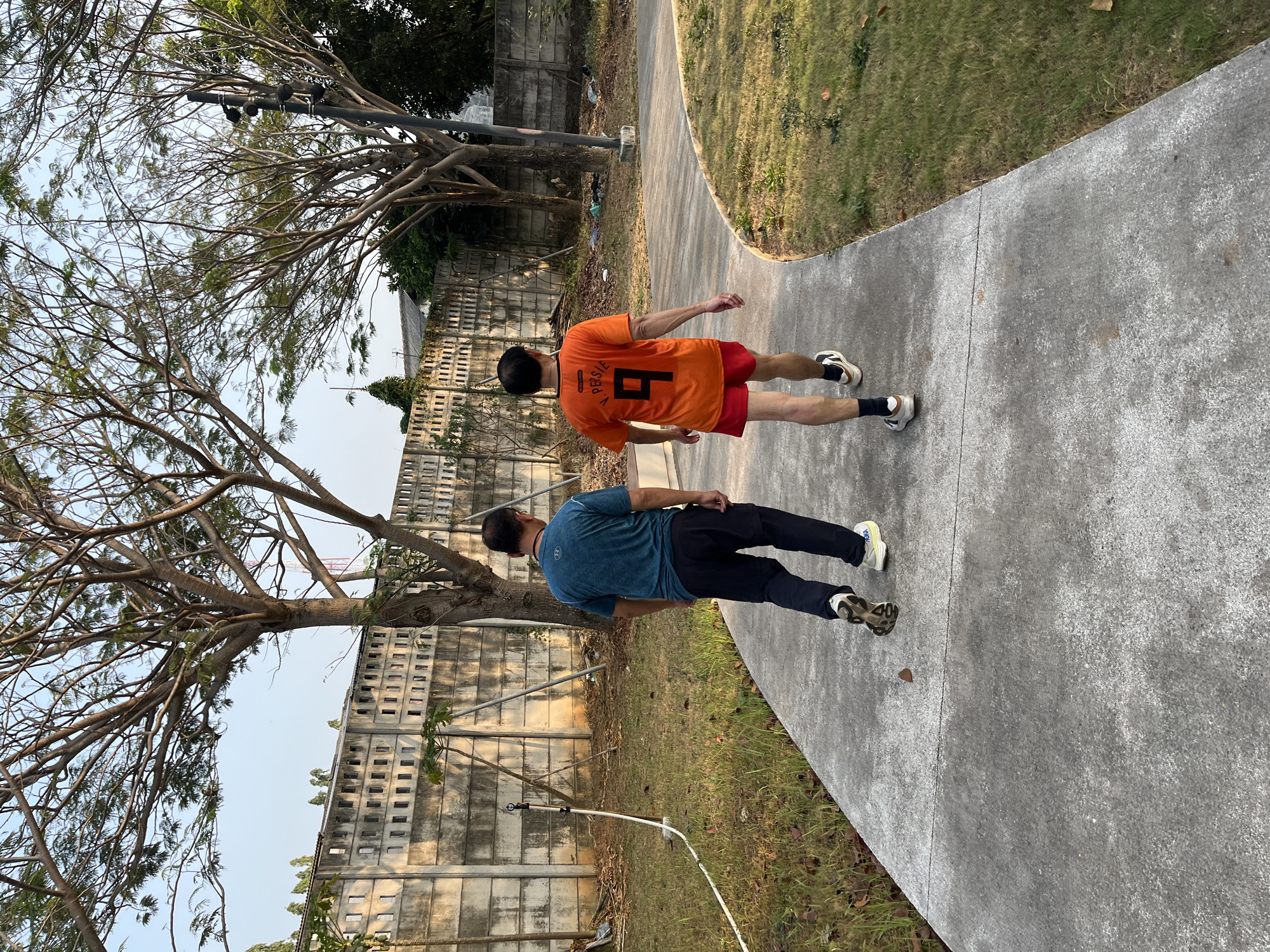 Two men walk away from the camera on a paved pathway in a park-like setting. The man on the left wears a teal t-shirt and dark pants. The man on the right wears an orange t-shirt with the number 9 and "V. PERSIE" printed on the back, and red shorts. Both men are wearing athletic shoes. The pathway is bordered by grass and trees. In the background, a concrete wall runs along the perimeter of the area. The sky is clear and blue. The scene appears to be outdoors during the daytime.