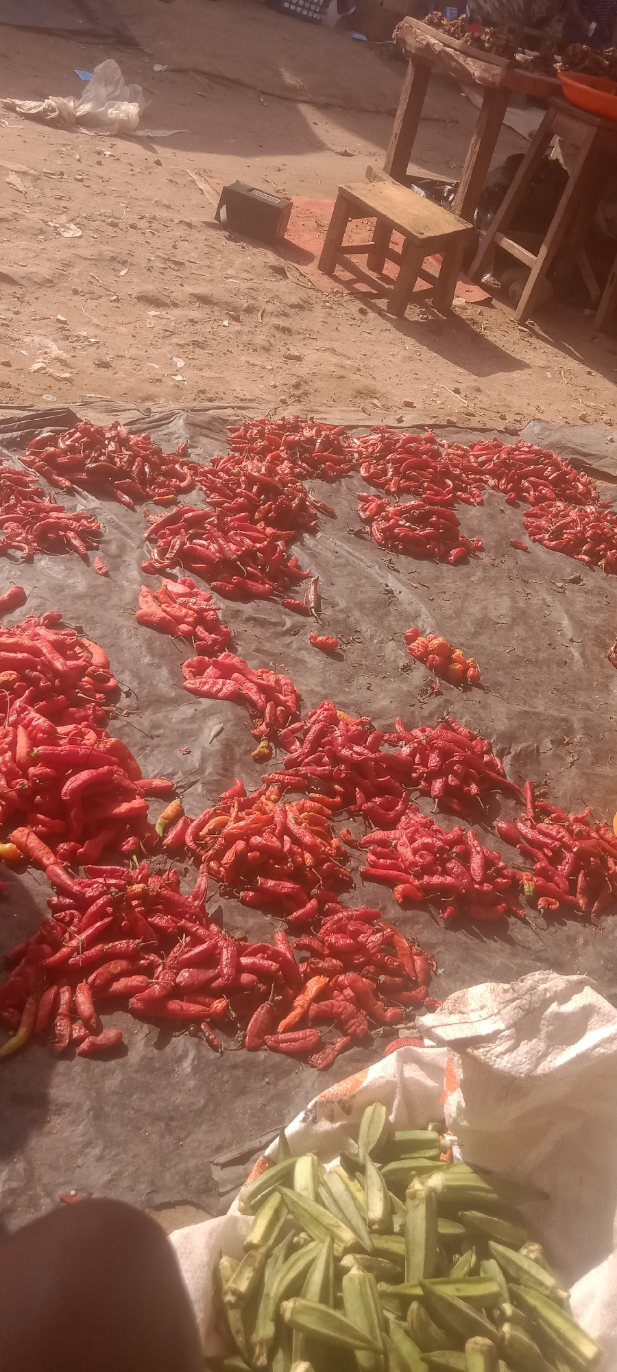 The image shows a street-level outdoor market scene in Umuahia, Nigeria. Red peppers are drying on a dark tarp covering the ground. A bag of okra is visible in the foreground. In the background are simple wooden tables and stools. The ground is sandy. There are no people visible.