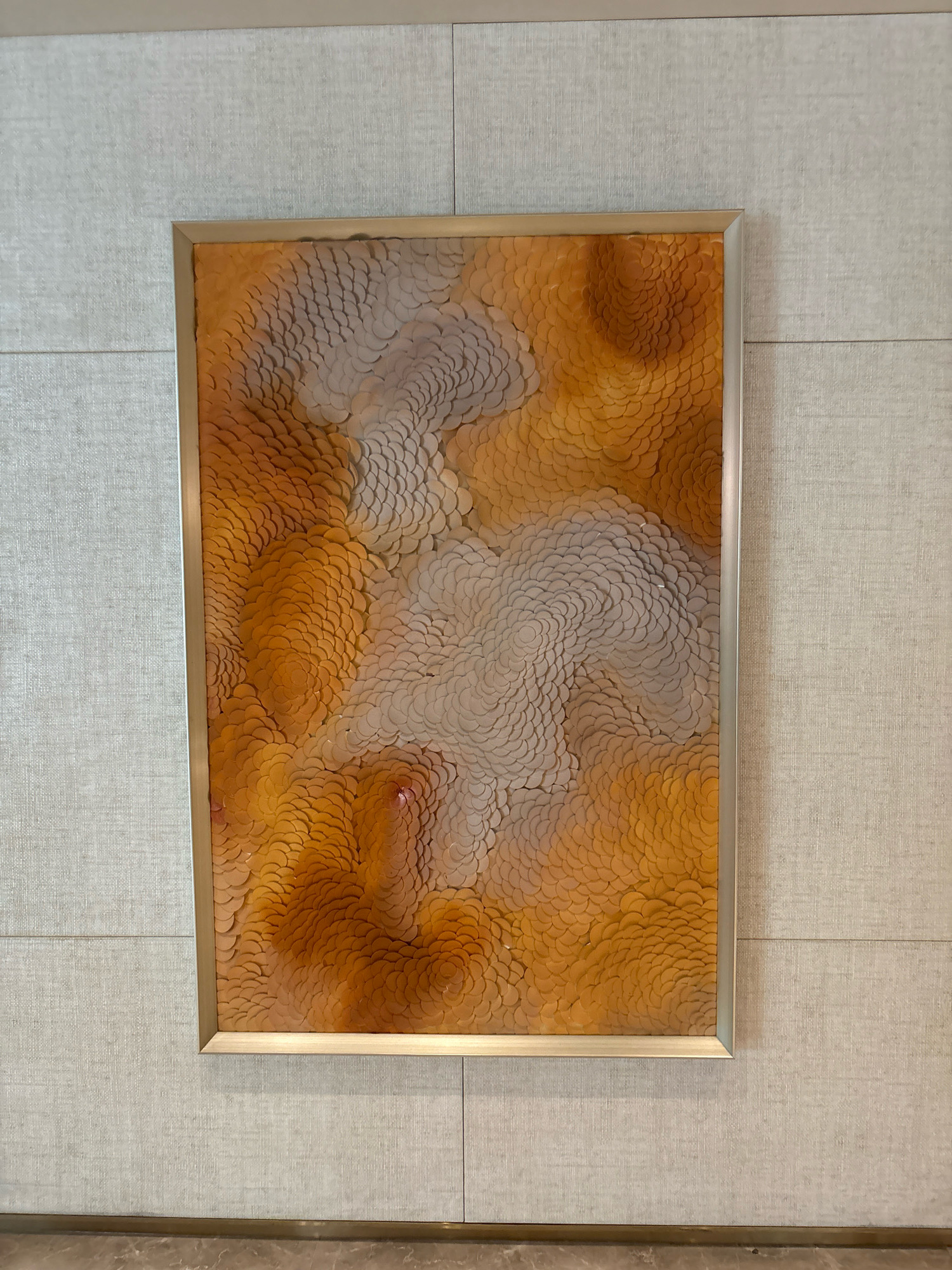 A framed artwork hangs on a textured, light-colored wall. The artwork is rectangular and features a pattern of overlapping circular or petal-like shapes in shades of orange and white, creating a textured, organic design. The frame is thin and metallic, appearing gold or brass. The wall is divided into horizontal panels with a subtle woven texture. Below the wall, a marble-like floor is visible. The scene is an interior shot, likely a home or a public space, and the location in Vagator, India is not discernible from the image itself. There are no people, actions, or interactions depicted.