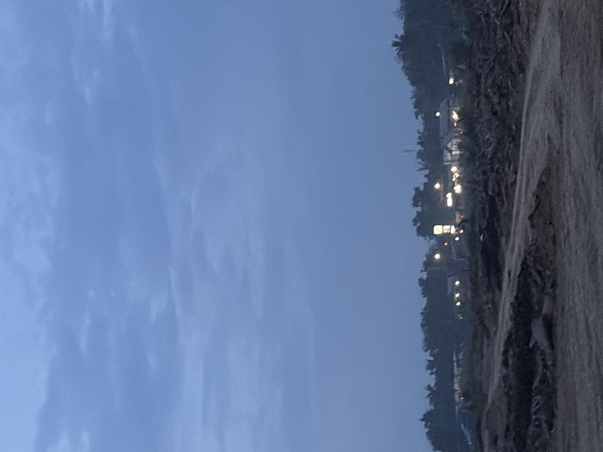 The photograph shows a view of Simpangempat, Indonesia, at dusk or dawn. The sky, occupying the majority of the image, is a pale blue with subtle cloud patterns. A strip of land runs along the right edge of the frame, consisting of sandy terrain or beach meeting vegetation, possibly trees. Lights are scattered among the vegetation, indicating the presence of buildings or streetlights.