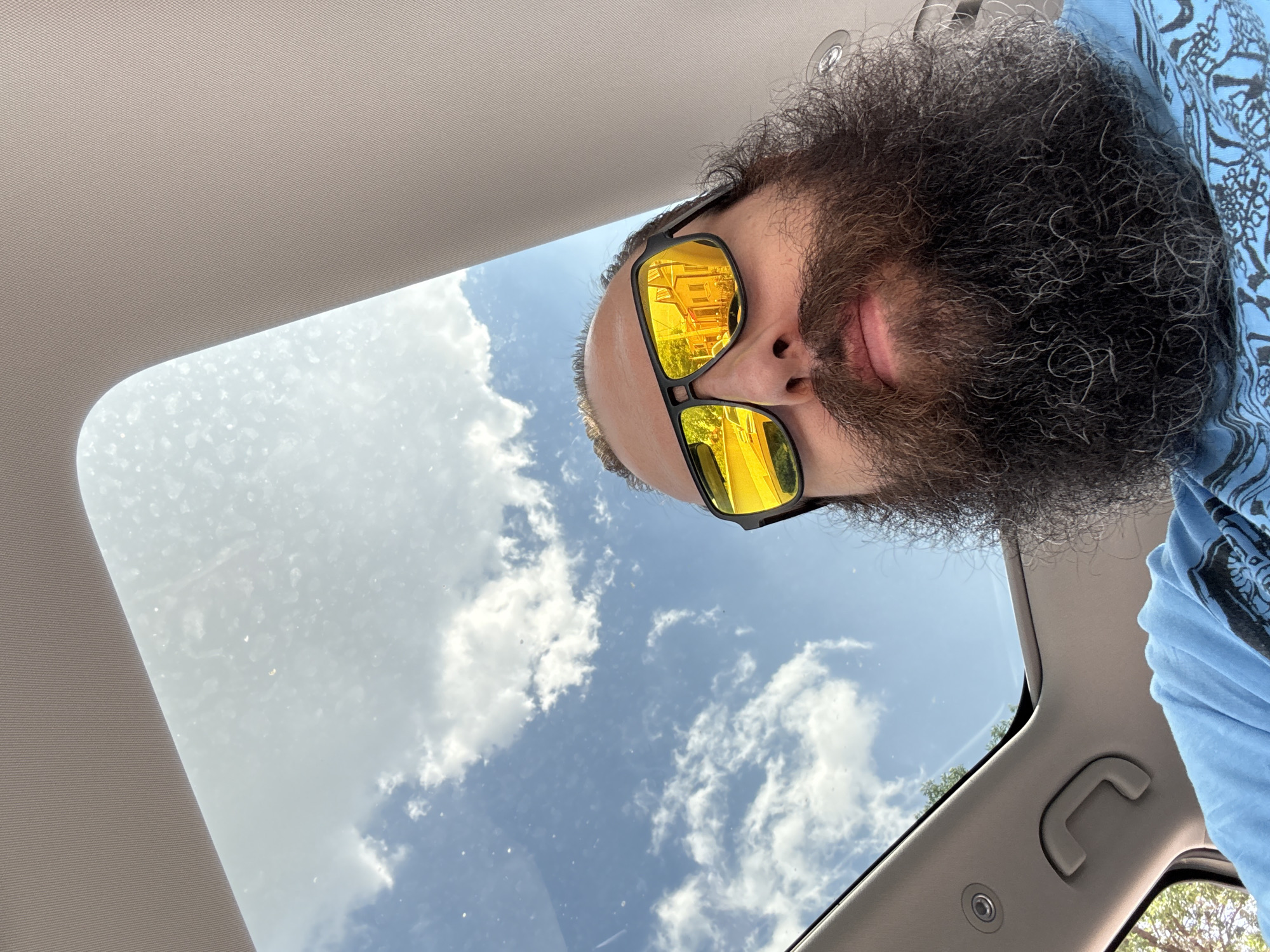 A man with a full beard is looking down from the inside of a car through a sunroof. He is wearing sunglasses that reflect a yellow building with several stories. The sky visible through the sunroof is blue with scattered white clouds. Part of the car's interior is visible, including the headliner and a grab handle. His shirt is blue and patterned. This cannot be placed in Tashkent, Uzbekistan.