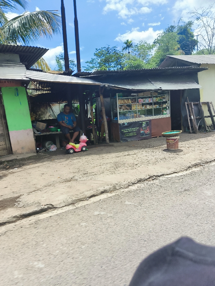 A man is sitting on a bench in front of a small shop in Cipatat, Indonesia, at coordinates -6.777 latitude and 107.437 longitude. The shop is a wooden structure with a corrugated metal roof and has a glass display case filled with various items, including bread and snacks. There is a small pink toy car in front of the man. The ground is dirt and gravel, and there is a paved road in the foreground. The sky is blue with white clouds. The man is wearing a blue shirt and dark pants, and is looking down at his phone. There are trees and bushes surrounding the shop. The man is not interacting with anyone.