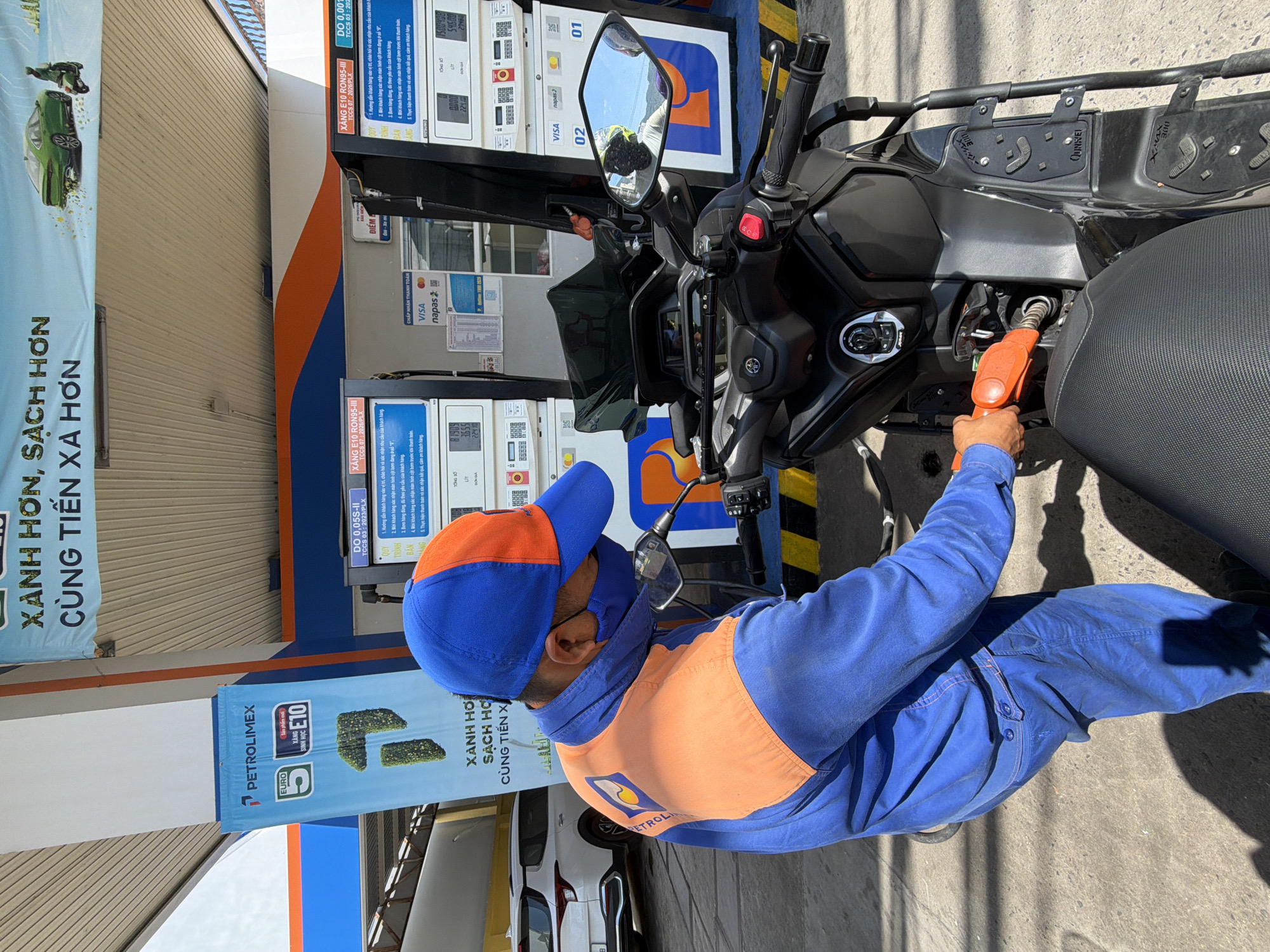 A person wearing a blue and orange uniform, a blue mask, and a blue and orange cap is fueling a black motorcycle at a Petrolimex gas station in Đại Lãnh, Vietnam. The person is holding an orange fuel nozzle and inserting it into the motorcycle's fuel tank. The gas station has a blue and white canopy, and there are gas pumps visible in the background. A white car is parked to the left of the frame. The scene is illuminated by natural daylight.