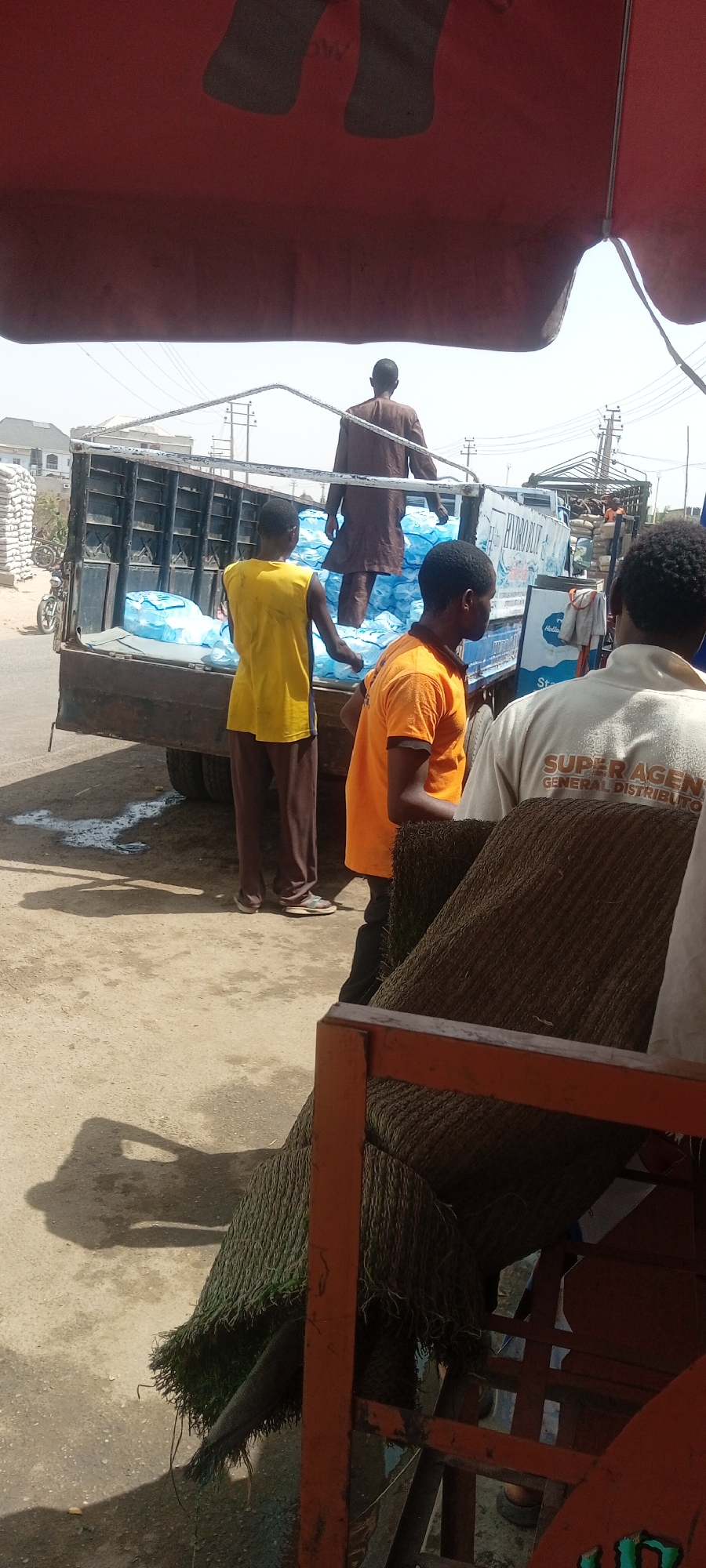 The image, taken during daylight in Aviation Quaters, Nigeria, depicts a scene of manual labor and commerce. A truck filled with blue plastic bags, likely containing water or another liquid, is being unloaded by three men. One man, wearing a brown robe, stands on top of the load in the truck bed. Another man, in a yellow tank top and brown pants, stands on the left side of the truck. A third man, wearing an orange t-shirt and black pants, is positioned on the right side of the truck bed, interacting with those unloading. A fourth man, wearing a white shirt with "SUPER AGENT GENERAL DISTRIBUTOR" printed on the back, stands on the far right, looking towards the scene.
In the foreground is what appears to be a partially obscured cart, possibly used for further transport of the goods, and the edge of a red awning overhead. Other buildings and a motorcycle are visible in the background, suggesting a semi-urban setting. Evidence of spillage or liquid discharge from the bags is apparent on the ground near the truck.