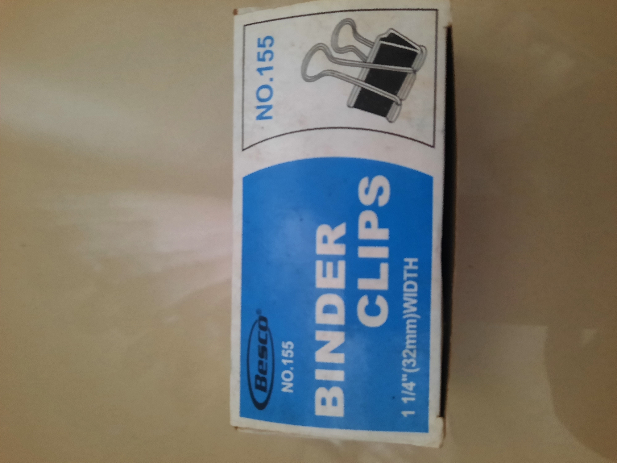 The image displays a box of "Besco" brand binder clips. The box is primarily blue and white, with prominent text indicating "NO.155", "BINDER CLIPS", and "1 1/4\"(32mm)WIDTH". A line drawing of two binder clips is also visible on the right side of the box. The box is placed on a light-colored, textured surface, likely a table or countertop, which appears to be indoors. The lighting suggests a natural light source, possibly from a window, creating subtle shadows and highlights. There is no visible indication of the city of Lhokseumawe, Indonesia, within the image itself.