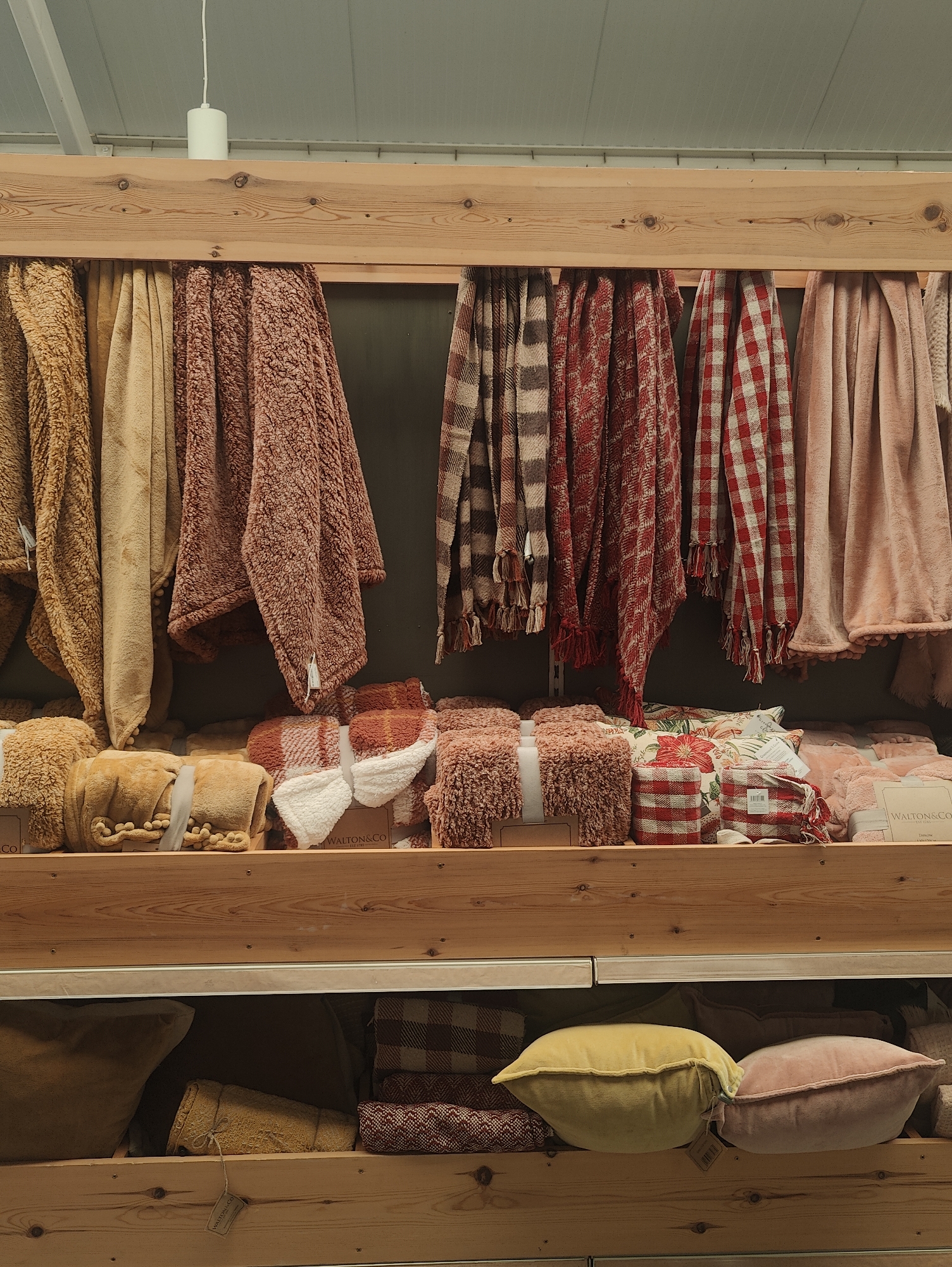 The image captures a scene inside a shop in Colchester, United Kingdom. The focus is on a wooden shelving unit filled with textiles, including throws and cushions. The top shelf showcases hanging throws in various colors and patterns, such as brown, beige, pink, plaid, and red gingham. Below the hanging throws are neatly stacked throws and cushions of similar colors and patterns, some adorned with tags and tied with ribbons or twine. The bottom shelf displays an assortment of cushions, including yellow and pink velvet pillows, along with other patterned textiles. The overall impression is that of a home goods or lifestyle store, specializing in soft furnishings. A white light fixture is visible on the ceiling above the shelves.