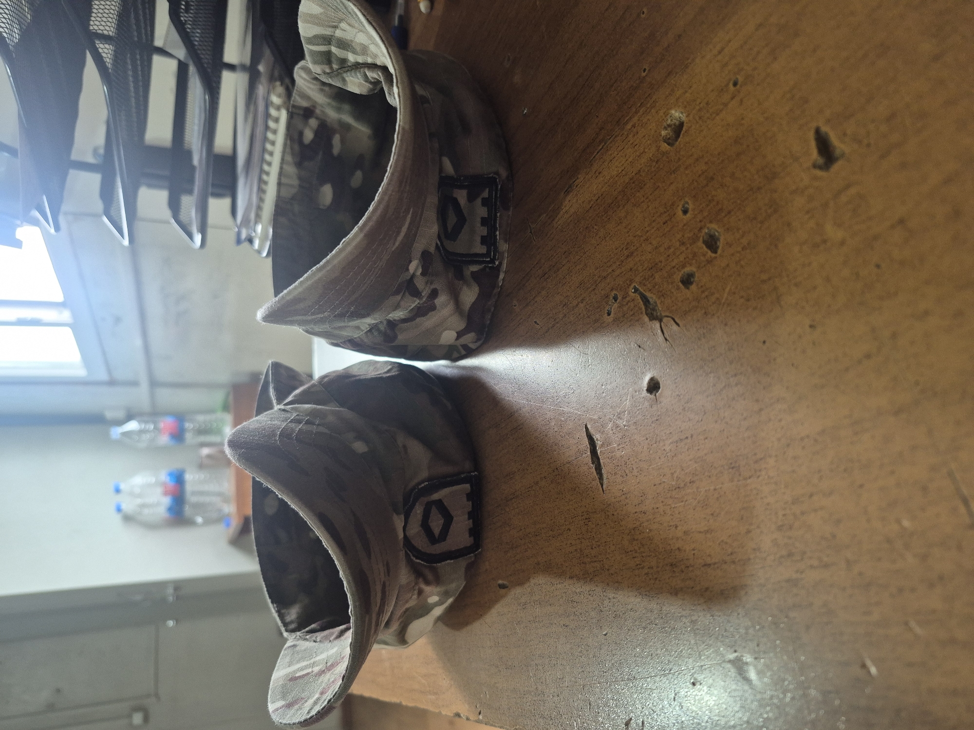 The image shows two camouflage caps placed on a wooden surface. Both caps have a rectangular patch with a diamond symbol on their sides. In the background, there is a clothes rack, a window, and some plastic water bottles. The wooden surface appears to be scratched and worn. The location in Tbilisi, Georgia, is not discernible from the given image.