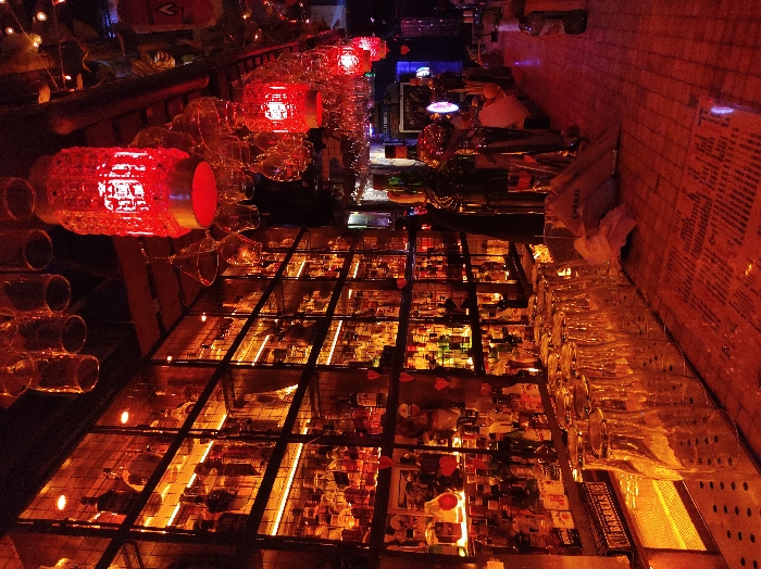 The image depicts the interior of a dimly lit bar, likely Openbaar Bar at Niceto Vega 5956, Palermo, Buenos Aires, Argentina, characterized by a dominant reddish-orange ambient light. The focal point is a large, floor-to-ceiling metal grid shelving unit on the left, densely stocked with hundreds of assorted bottles of alcoholic beverages, backlit by horizontal LED strips. Small red heart-shaped decorations are affixed to the lower sections of the shelving. Above the bar, numerous clear stemmed glasses and tumblers hang inverted from racks, reflecting the red light. Several ornate, cylindrical red lanterns suspended from the ceiling serve as primary light sources. In the foreground, a wooden bar counter extends rightwards, featuring a neat row of inverted clear tumblers on a metallic drain mat. Further along the counter are various liquor bottles, metallic bar tools, and a stack of white napkins, one partially displaying "OPENBAAR." Printed menus or placemats lie flat on the counter surface. An indistinct dark figure is partially visible behind the bar counter on the right, near beer taps or dispensing equipment. No other individuals are clearly visible.